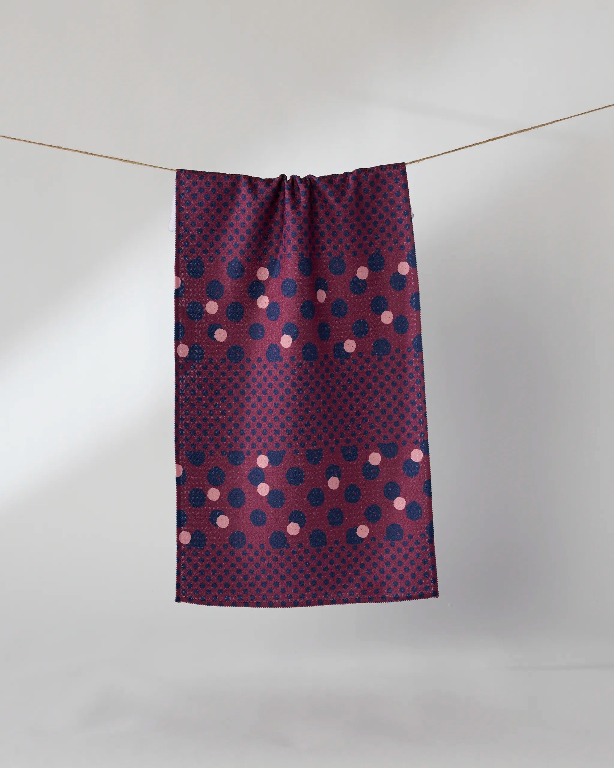 The Geometry Mulberry Dot bar towel features a navy and burgundy polka dot pattern with pink and purple accents. Made from recycled materials, it hangs on a string against a plain light gray background.
