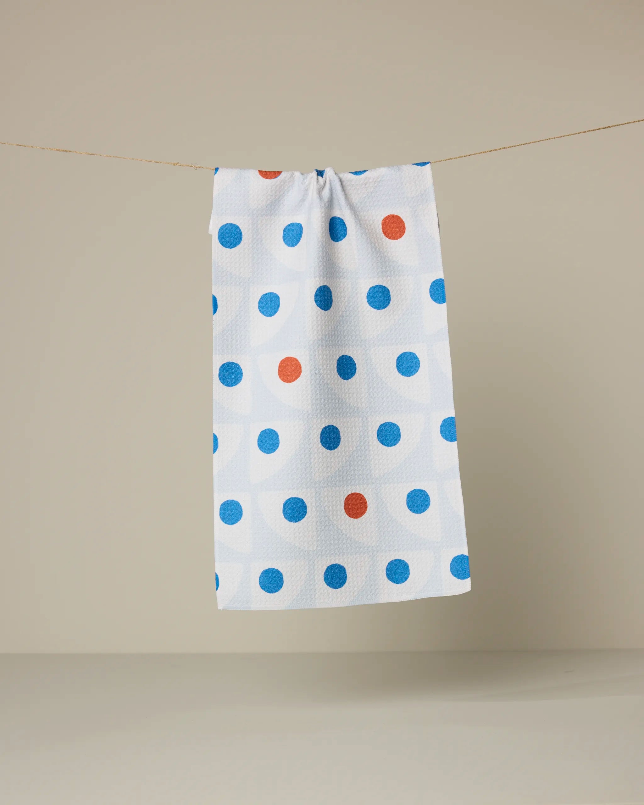 The Geometry Nautical Eye towel, featuring blue and orange polka dots on white, hangs from a thin string against a plain beige background.