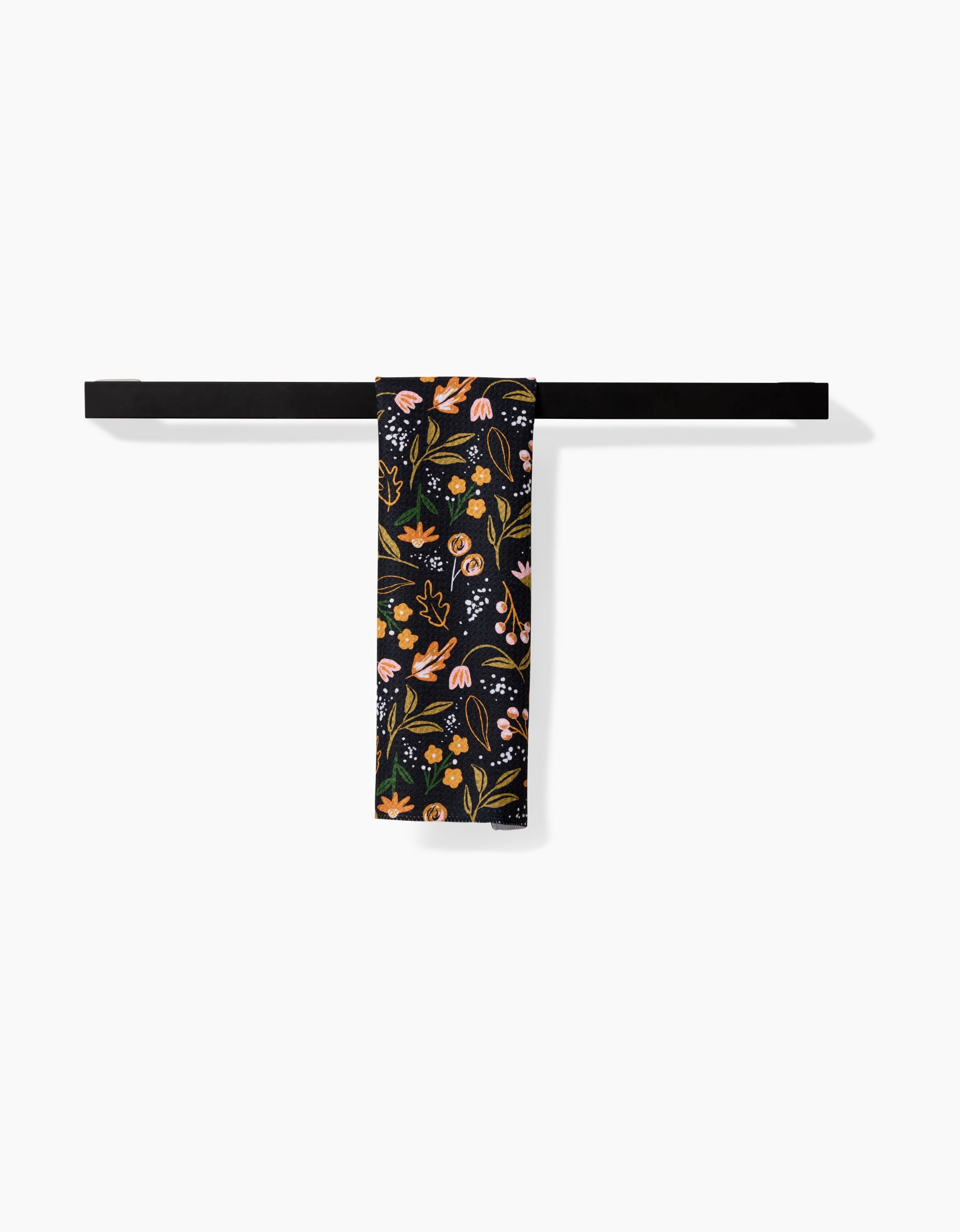 The Megan Roy Nighttime Botanical black bar towel, featuring vibrant floral and mushroom designs on Geoweave fabric, hangs neatly folded over a simple black towel bar against a white background.