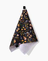The Megan Roy Nighttime Botanical bar towel features a colorful floral pattern in pink, yellow, and green on black, hangs from a black loop, and is made of Geoweave fabric with a white textured underside for absorbency and durability.