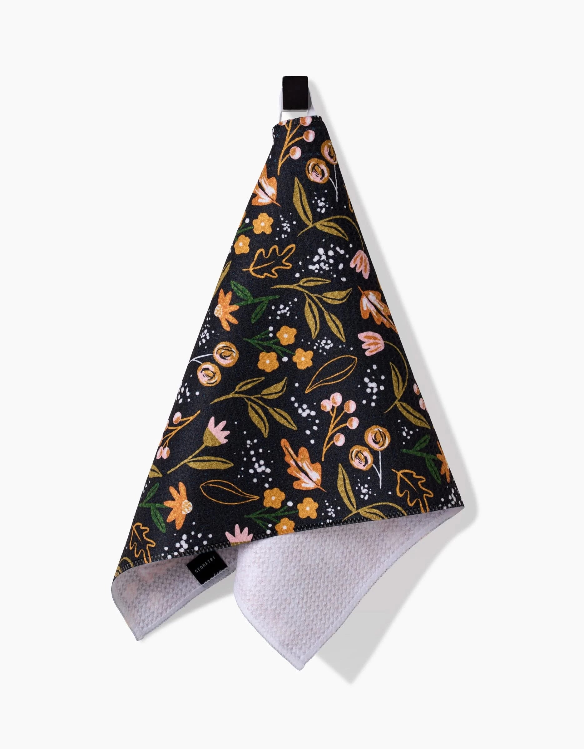 The Megan Roy Nighttime Botanical bar towel features a colorful floral pattern in pink, yellow, and green on black, hangs from a black loop, and is made of Geoweave fabric with a white textured underside for absorbency and durability.