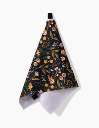 The Megan Roy Nighttime Botanical bar towel features a colorful floral pattern in pink, yellow, and green on black, hangs from a black loop, and is made of Geoweave fabric with a white textured underside for absorbency and durability.