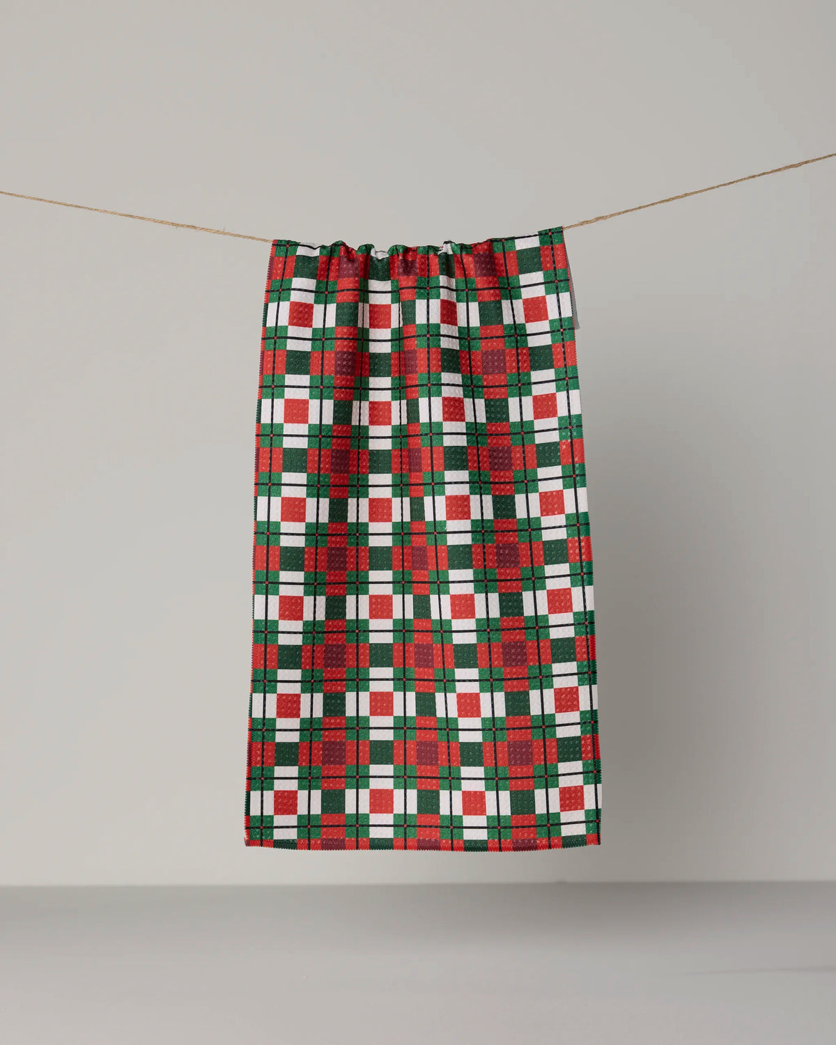 A Geometry Noel Plaid tea towel, crafted from Geoweave™ fabric and featuring a red, green, and white plaid pattern, hangs on a clothesline against a plain light gray background.