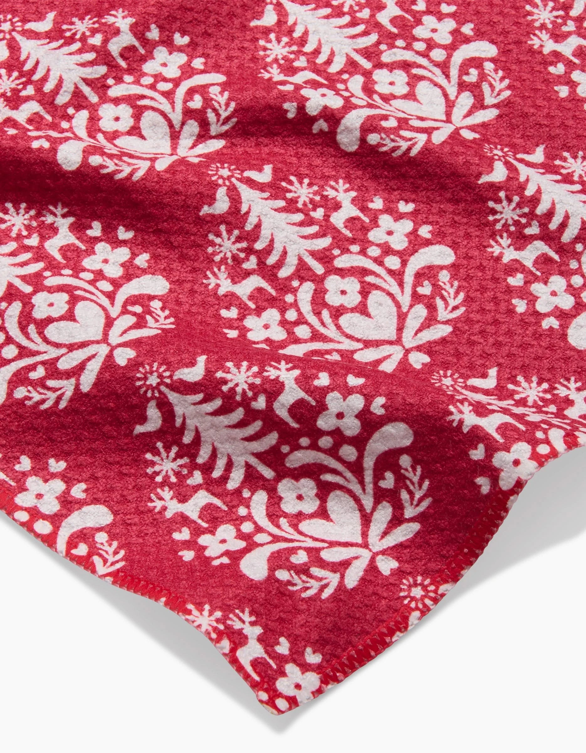 A close-up of Kayla Ann’s Nordic Holiday Stamp Red bar towel shows its recycled fabric with a white folk-inspired pattern—flowers, hearts, birds, and trees—plus a wavy edge and slightly textured surface.