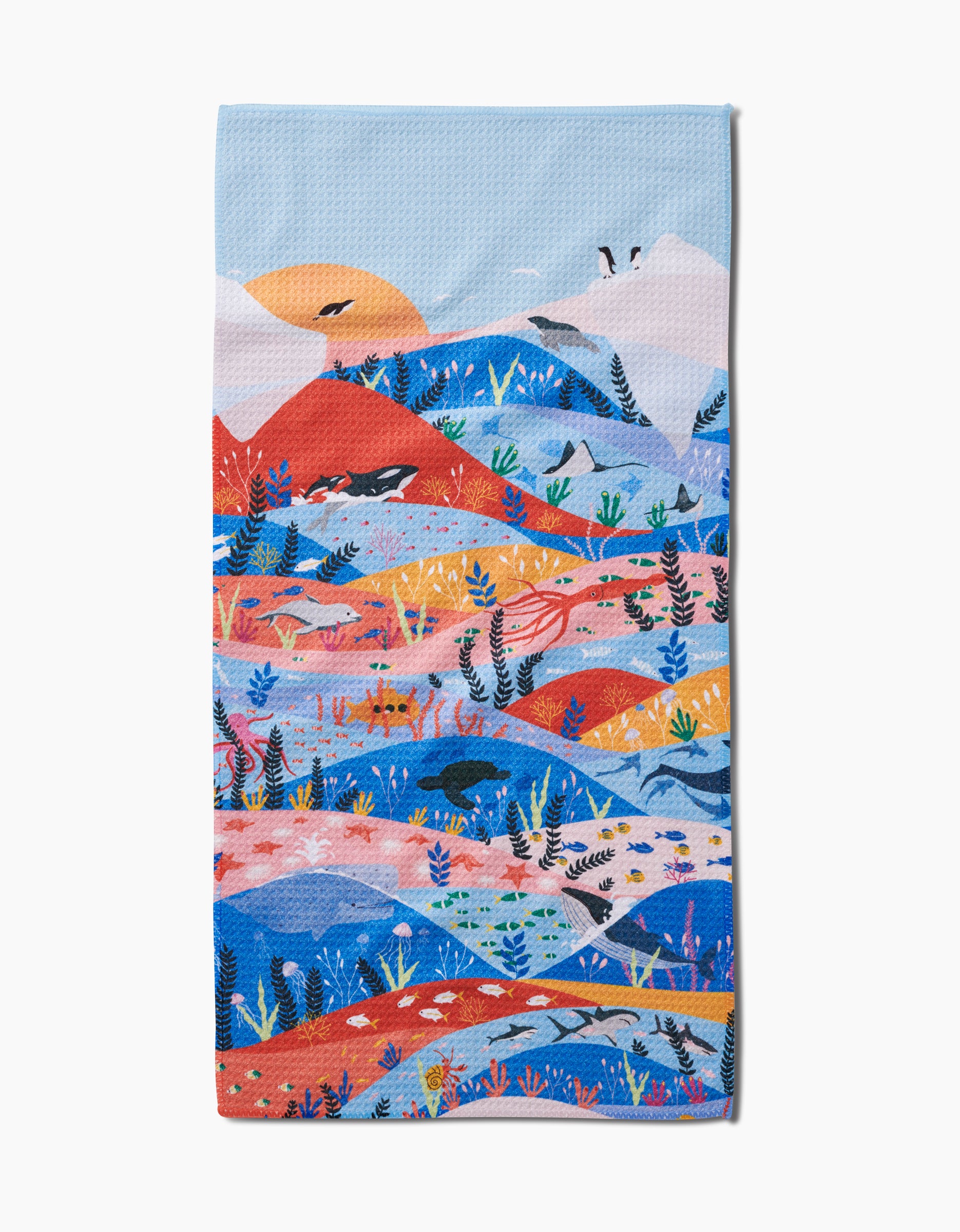 The Oceans bar towel by Ceyda Alasar is made from recycled materials and showcases a vibrant ocean scene with waves, marine life, plants, and a sunset in blue, orange, and red tones.