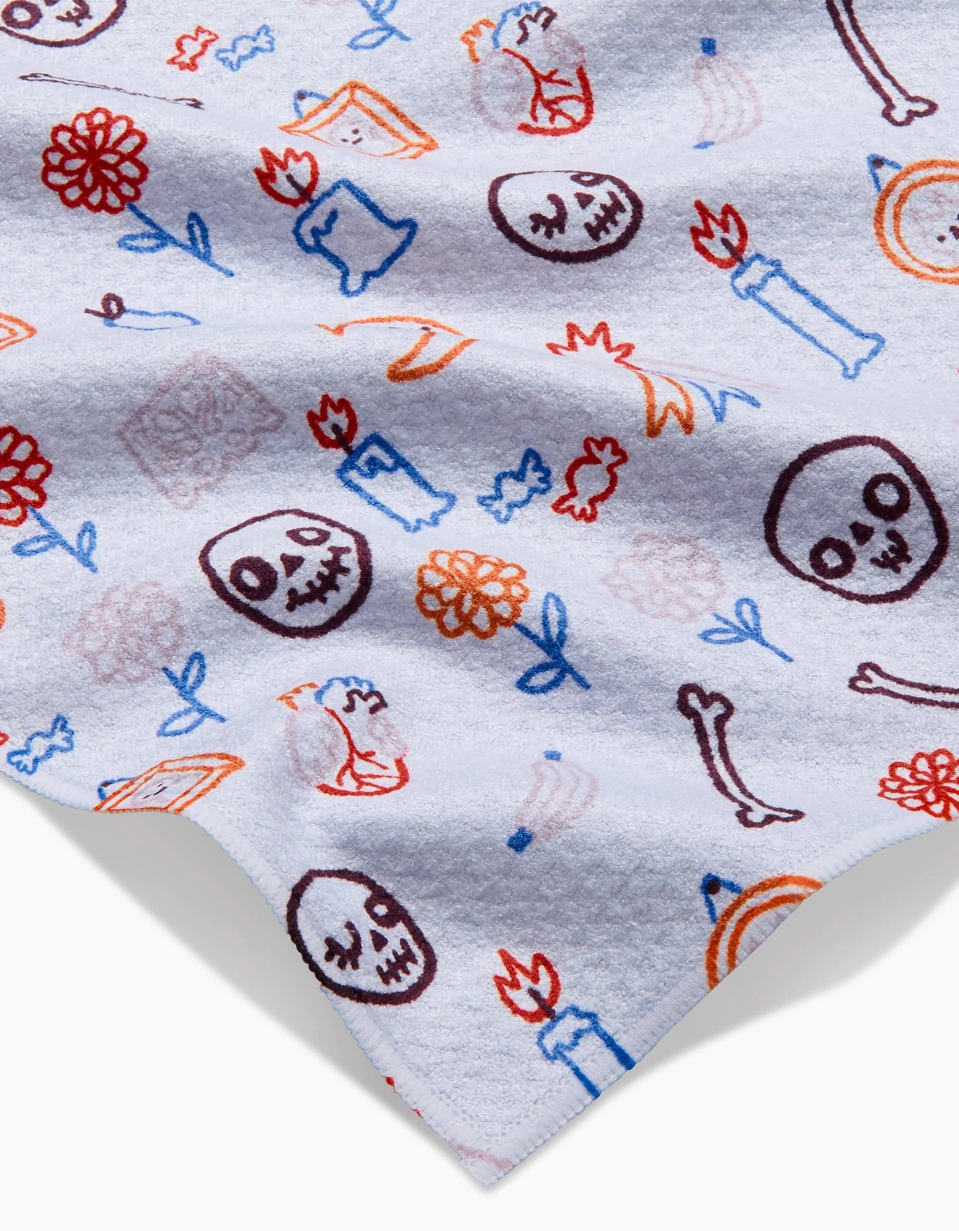 The Offerings Bar Towel by So Lazo features colorful, hand-drawn skulls, flowers, candles, frames, and bones in red, blue, and orange scattered on white Geoweave fabric.