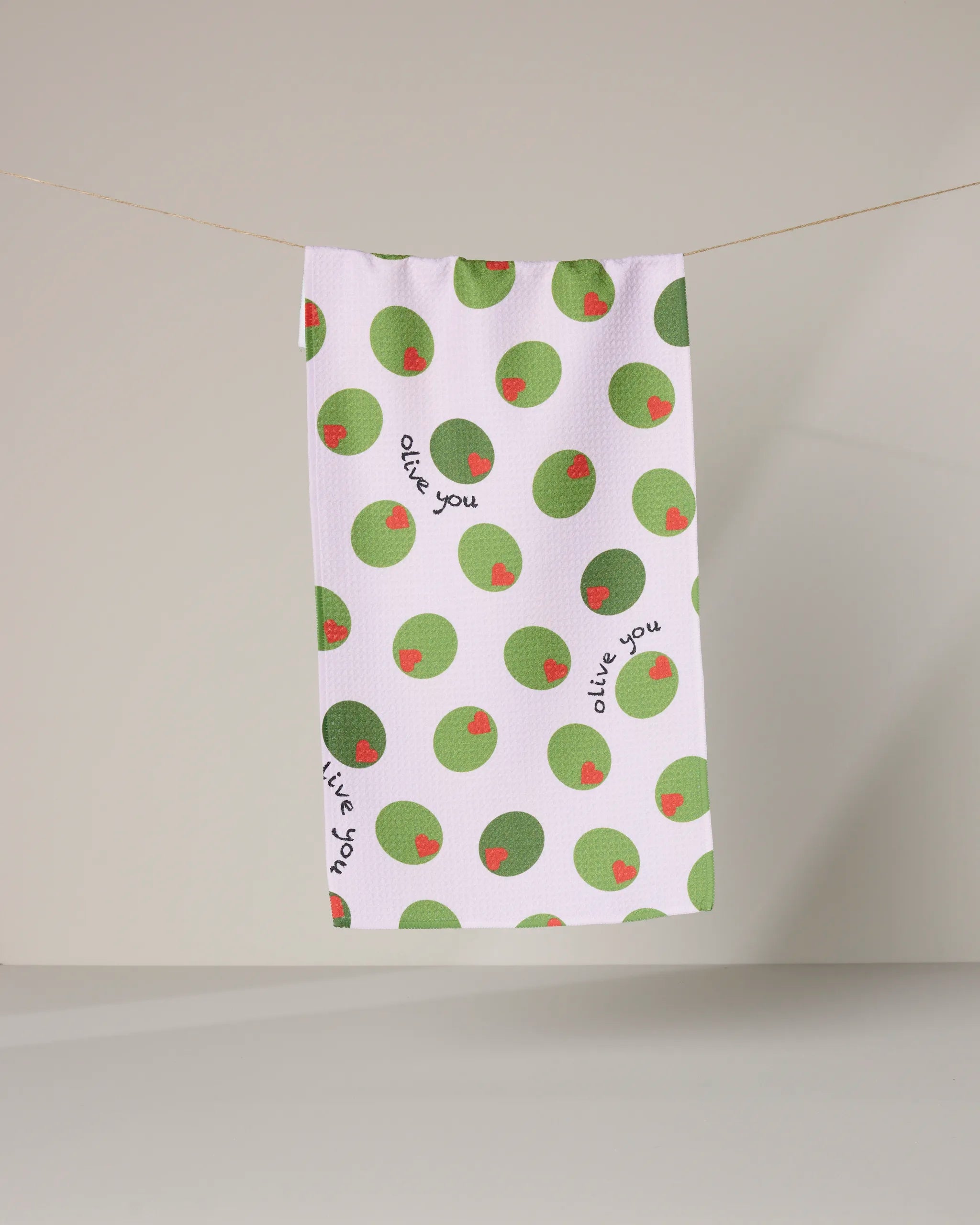 The Olive You towel by Geometry, featuring green olive illustrations and the phrase "olive you," hangs from a string against a plain background.