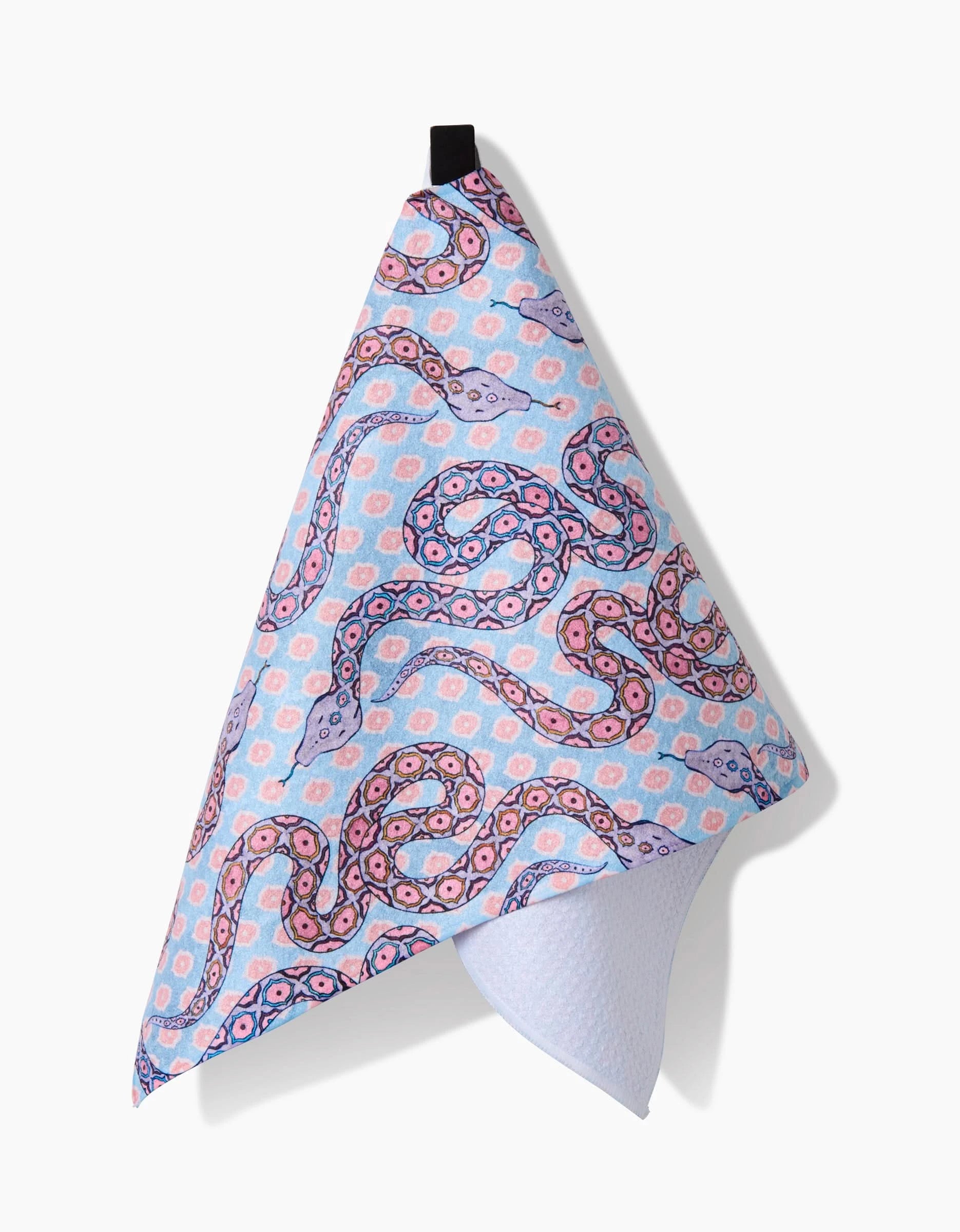 The Ophidia bar towel by Maayan Alper-Swan features a pink and purple snake motif on light blue, with a subtle hexagonal background, made from ultra-absorbent Geoweave fabric and hanging from a black loop against a white backdrop.