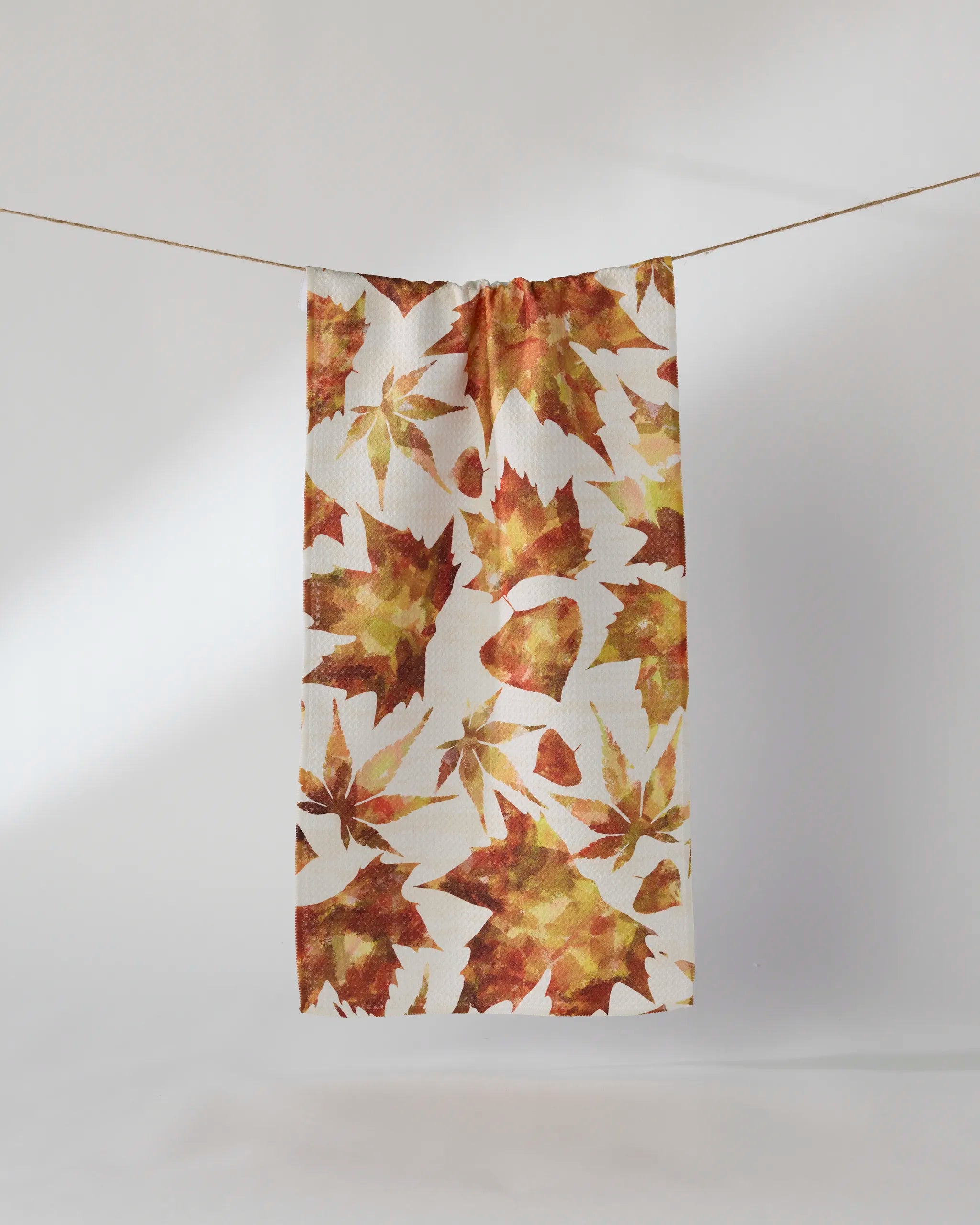 The Geometry Painted Fall towel, featuring an ultra-absorbent white fabric with orange, yellow, and brown autumn leaf prints, hangs on a clothesline against a plain light backdrop.