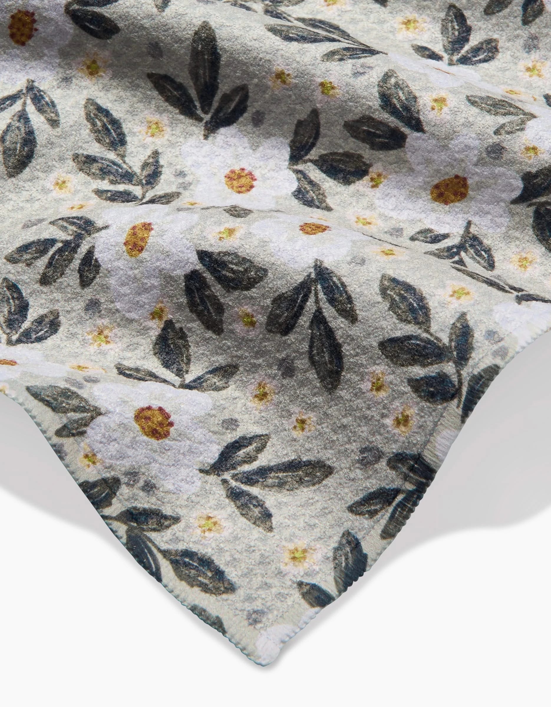 A close-up of Teri the Butter’s Painted Spring Flowers Geoweave bar towel, showcasing a soft fabric with white flowers, yellow centers, and dark green leaves on a light background. The corner edge appears at the bottom right.