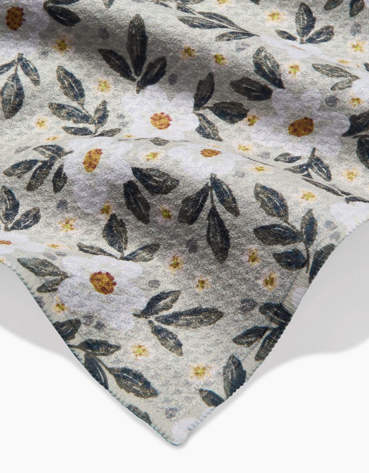 A close-up of Teri the Butter’s Painted Spring Flowers Geoweave bar towel, showcasing a soft fabric with white flowers, yellow centers, and dark green leaves on a light background. The corner edge appears at the bottom right.