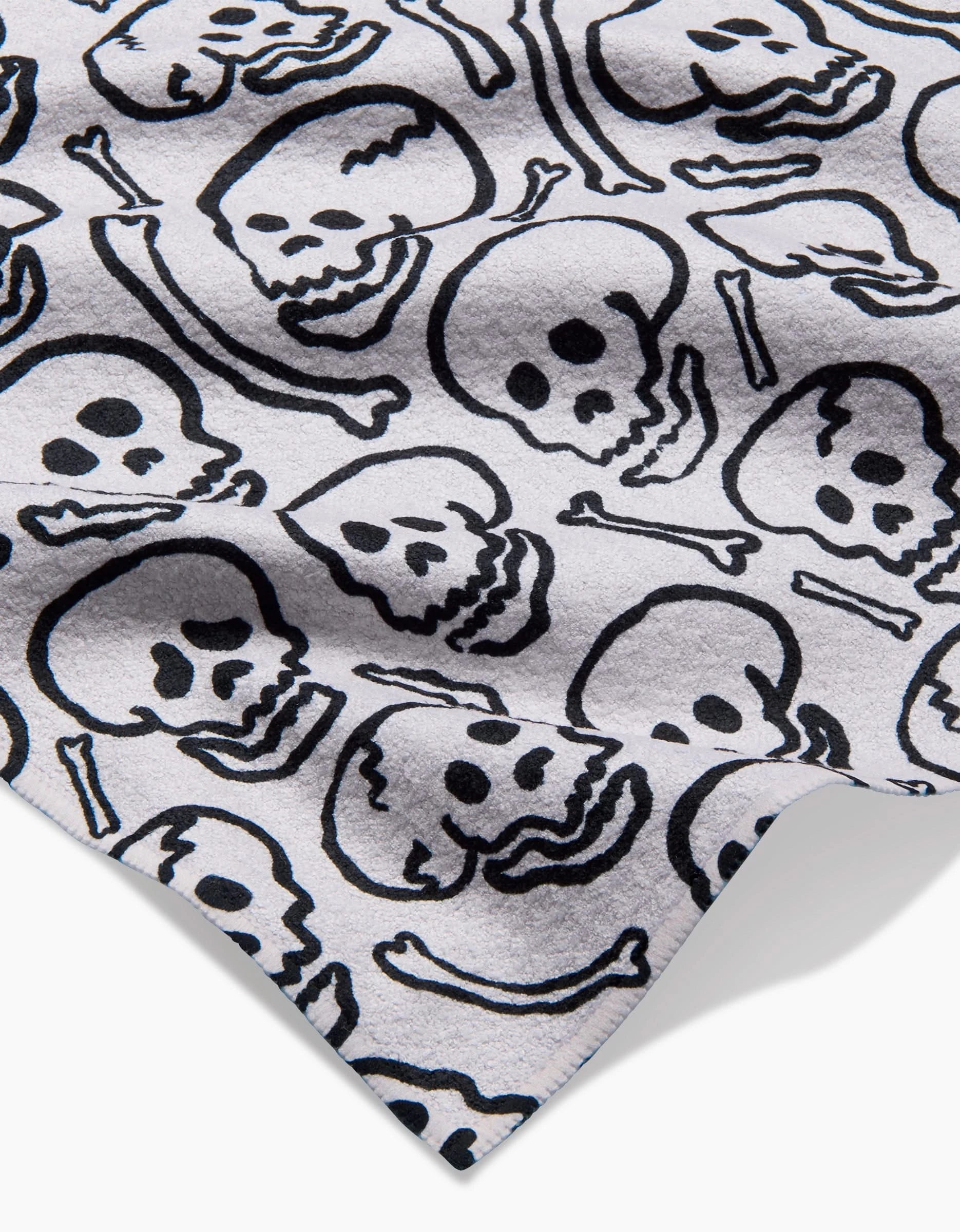 A close-up of Kayla Ann’s Painterly Skulls towel, featuring ultra-absorbent white fabric with a black, cartoon-style skull and bone pattern scattered across the surface.