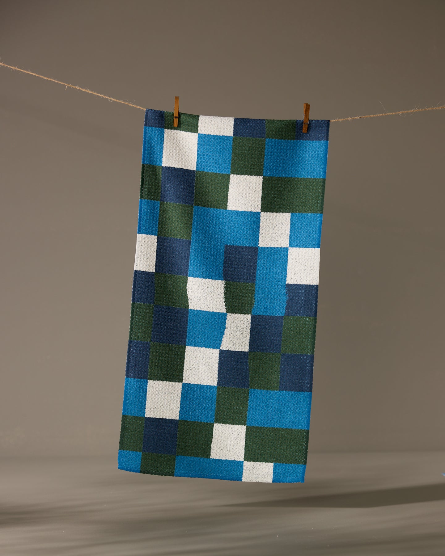 The Geometry Patchpoint towel features a rectangular shape and an ultra-absorbent design with a blue, green, white, and dark blue geometric square pattern, shown hanging from a clothesline with clothespins against a neutral background.