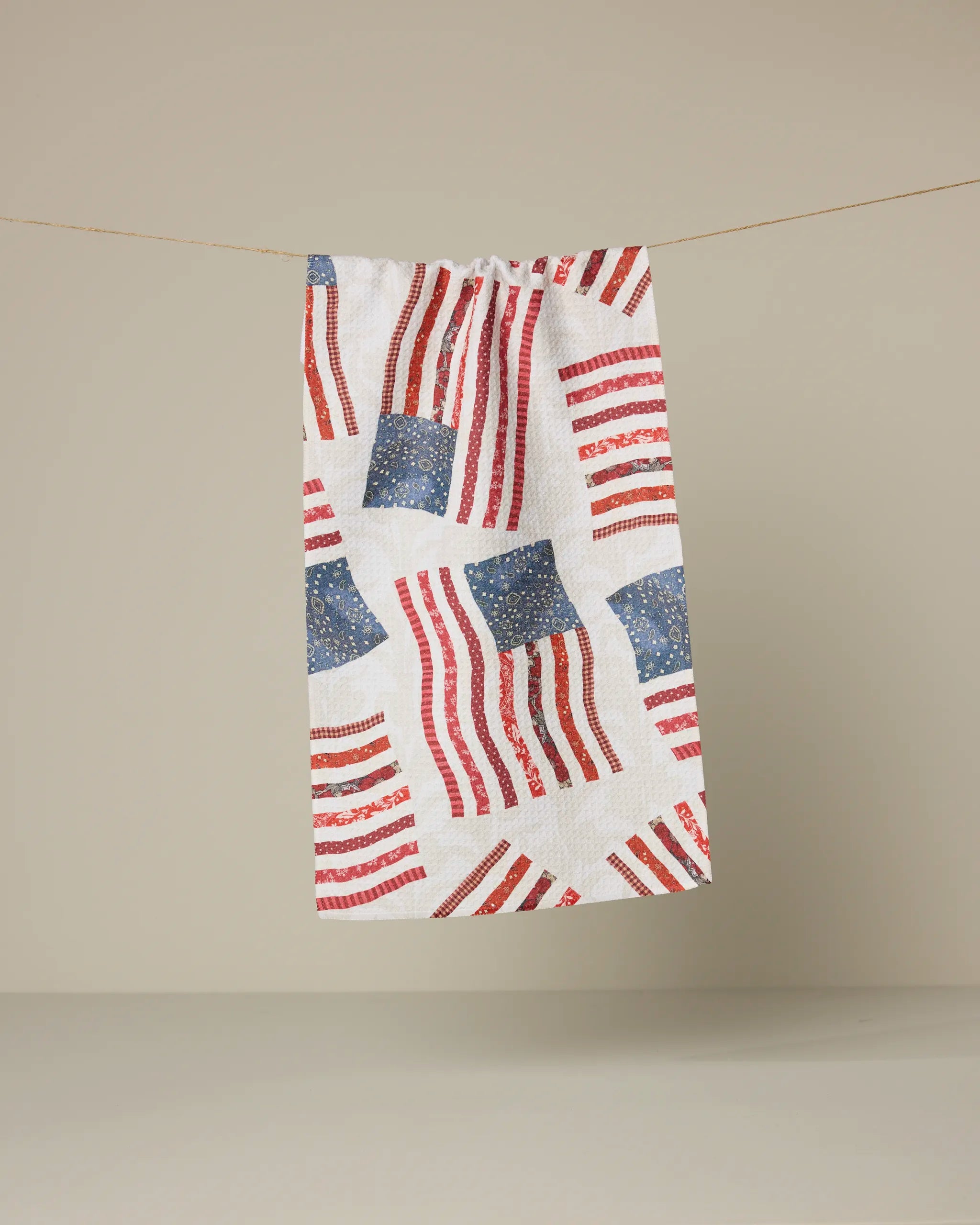 The Patchwork Pride kitchen towel by Geometry, made from recycled materials, features a repeating red, white, and blue American flag pattern and hangs against a plain beige background.
