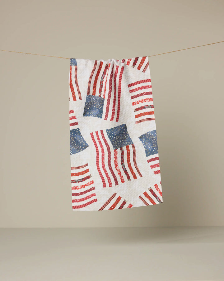 The Patchwork Pride kitchen towel by Geometry, made from recycled materials, features a repeating red, white, and blue American flag pattern and hangs against a plain beige background.