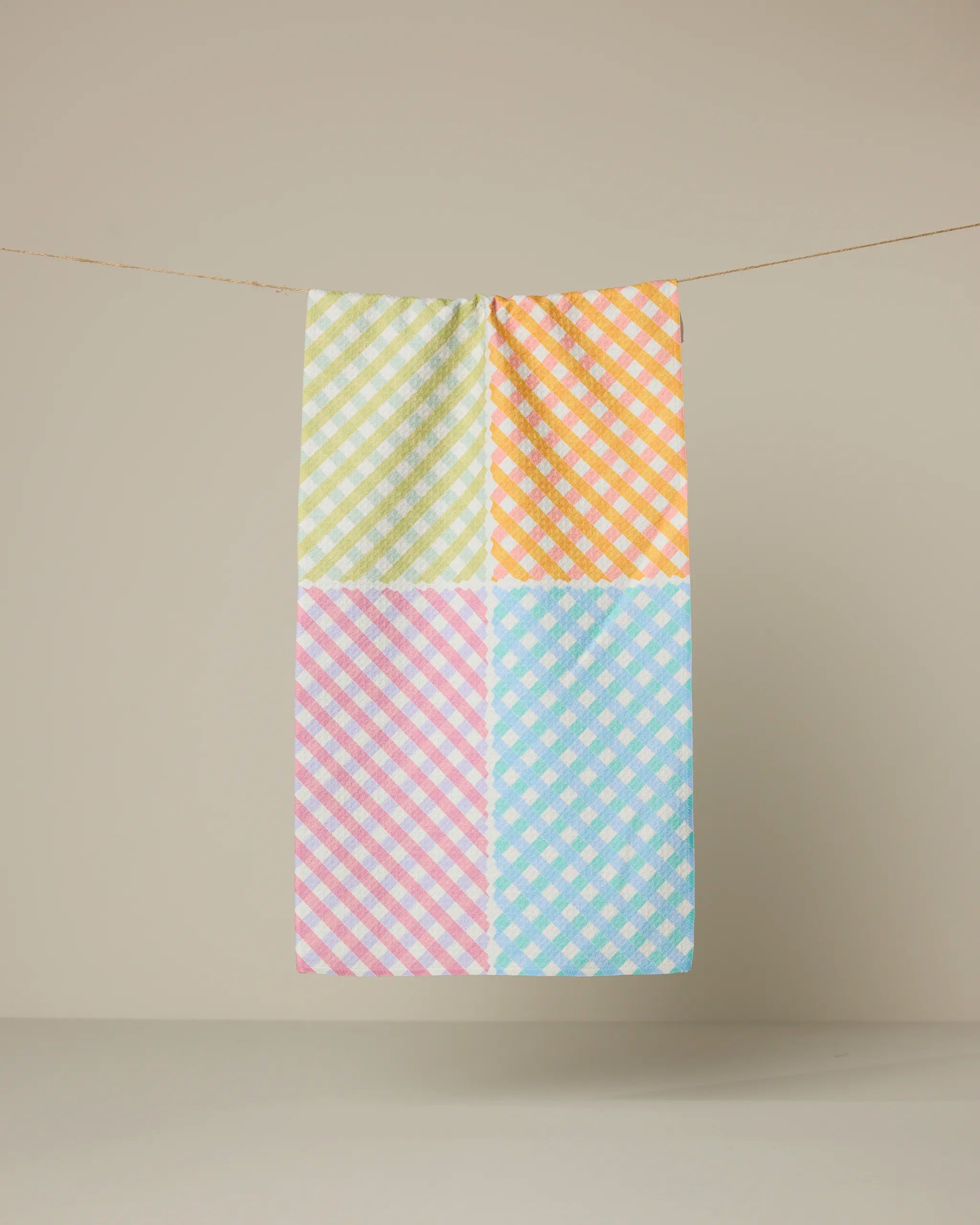 The Geometry Patio Check recycled kitchen towel, featuring a checkered pattern in green, orange, pink, and blue pastels, hangs from a string against a plain beige background.