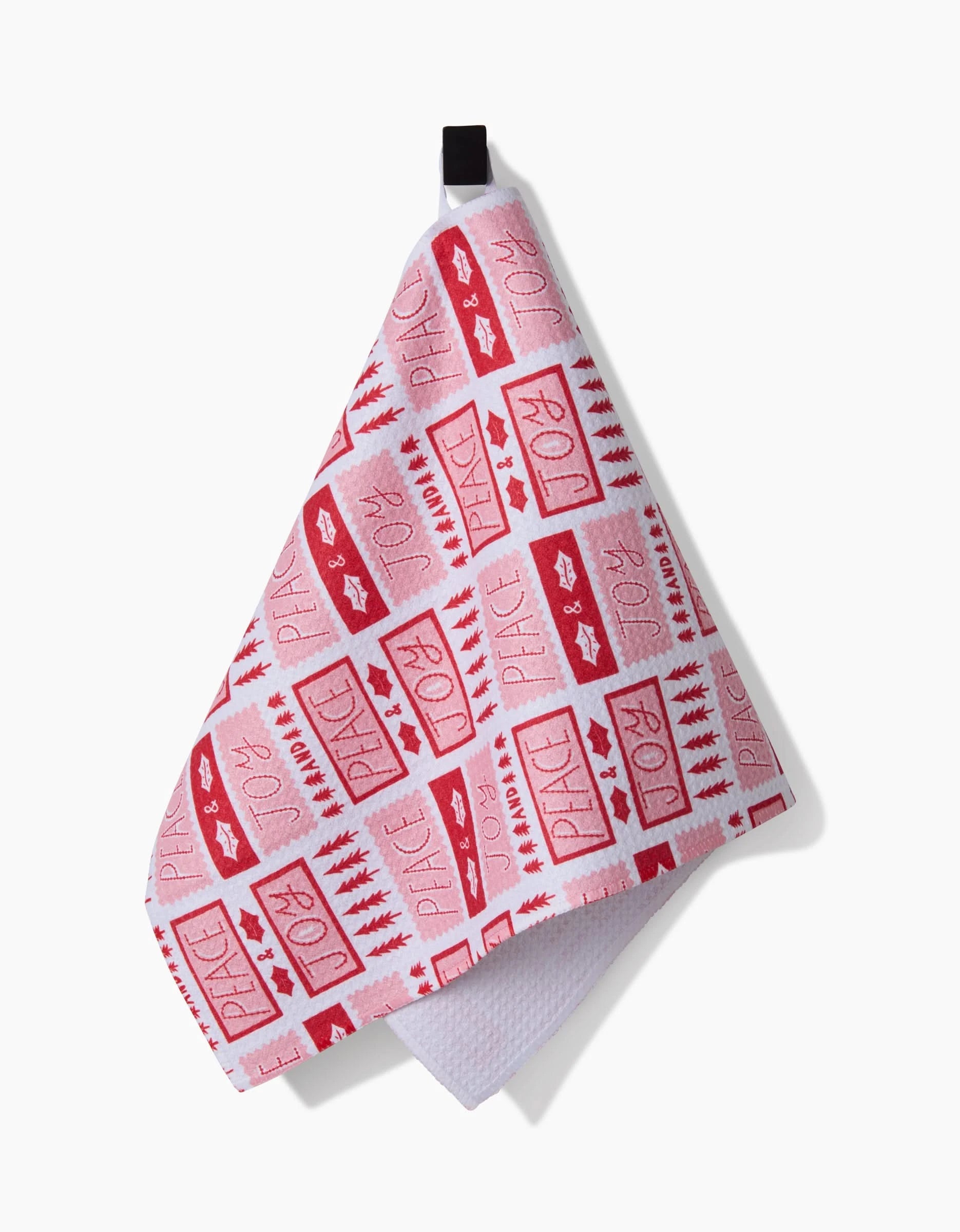 The Roxy Marj Peace Joy Stamps bar towel is ultra-absorbent and white, decorated with red stars, rectangles, Christmas trees, and festive words like JOY and PEACE. It hangs by a black loop.