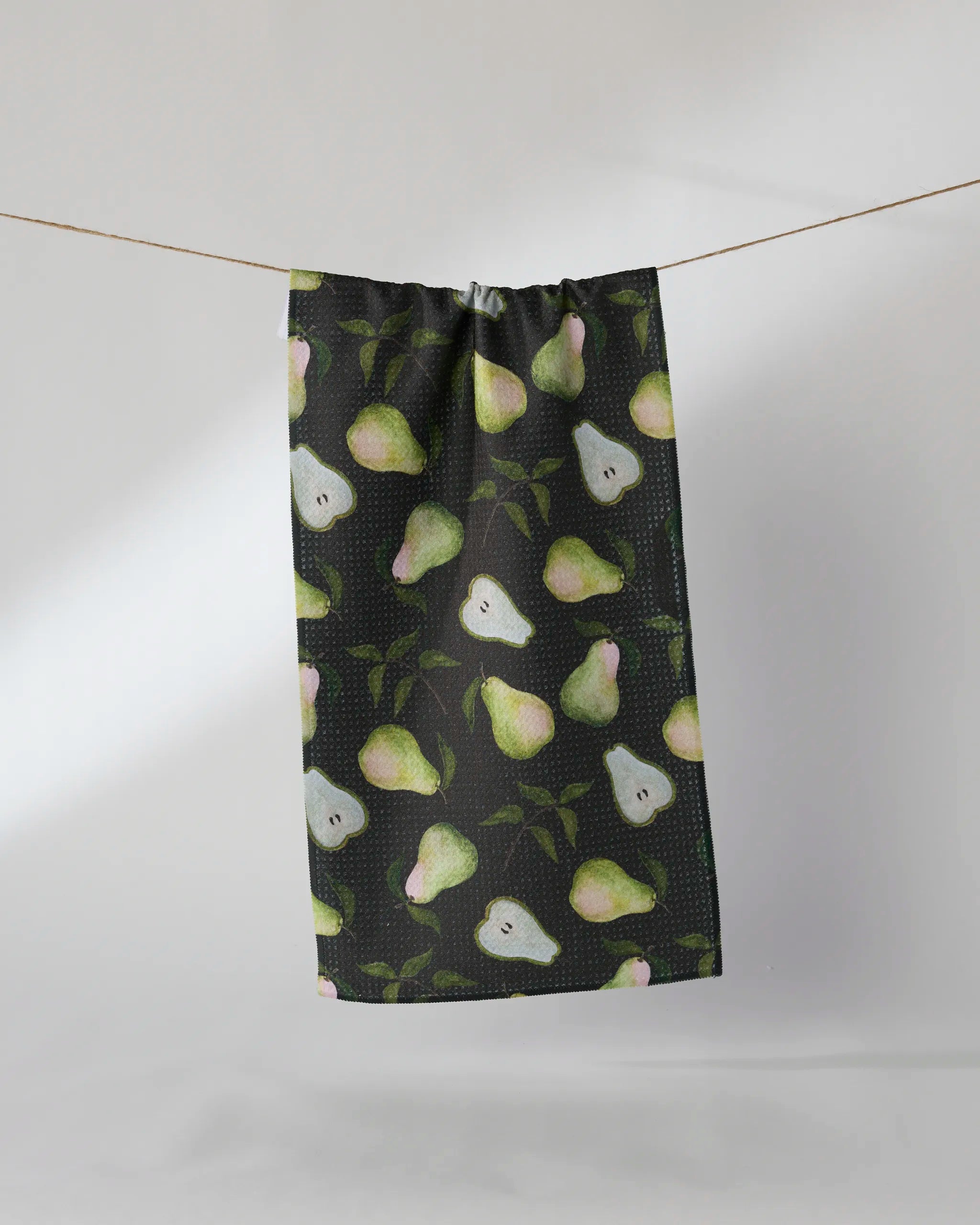 The Geometry Pearwood towel, ultra-absorbent and dark, features whole and sliced green pears with leafy accents, displayed hanging on a clothesline against a simple light background.