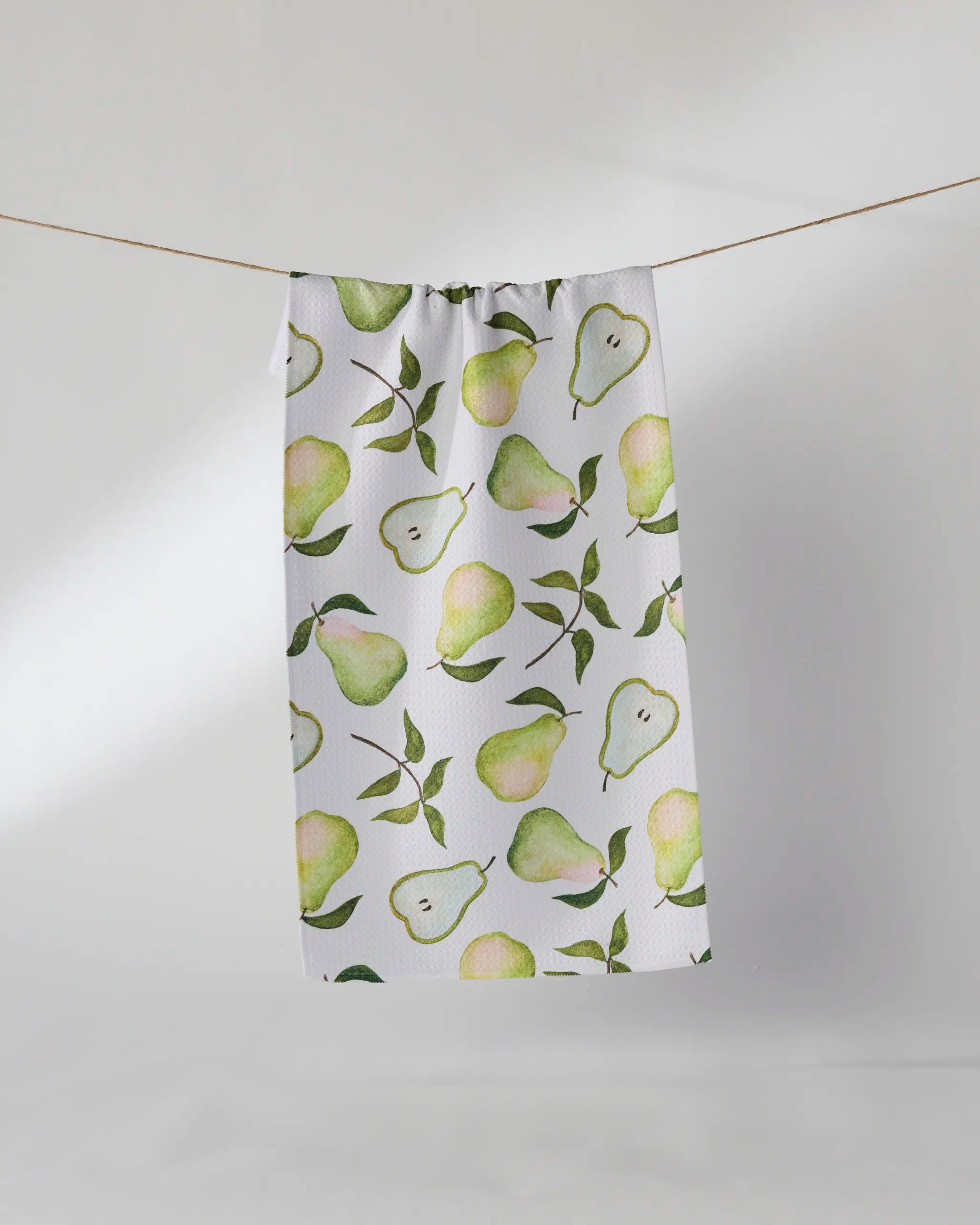 The Pearwood bar towel by Geometry, ultra-absorbent and white with a green pear and leaf pattern, hangs on a clothesline against a plain light background.
