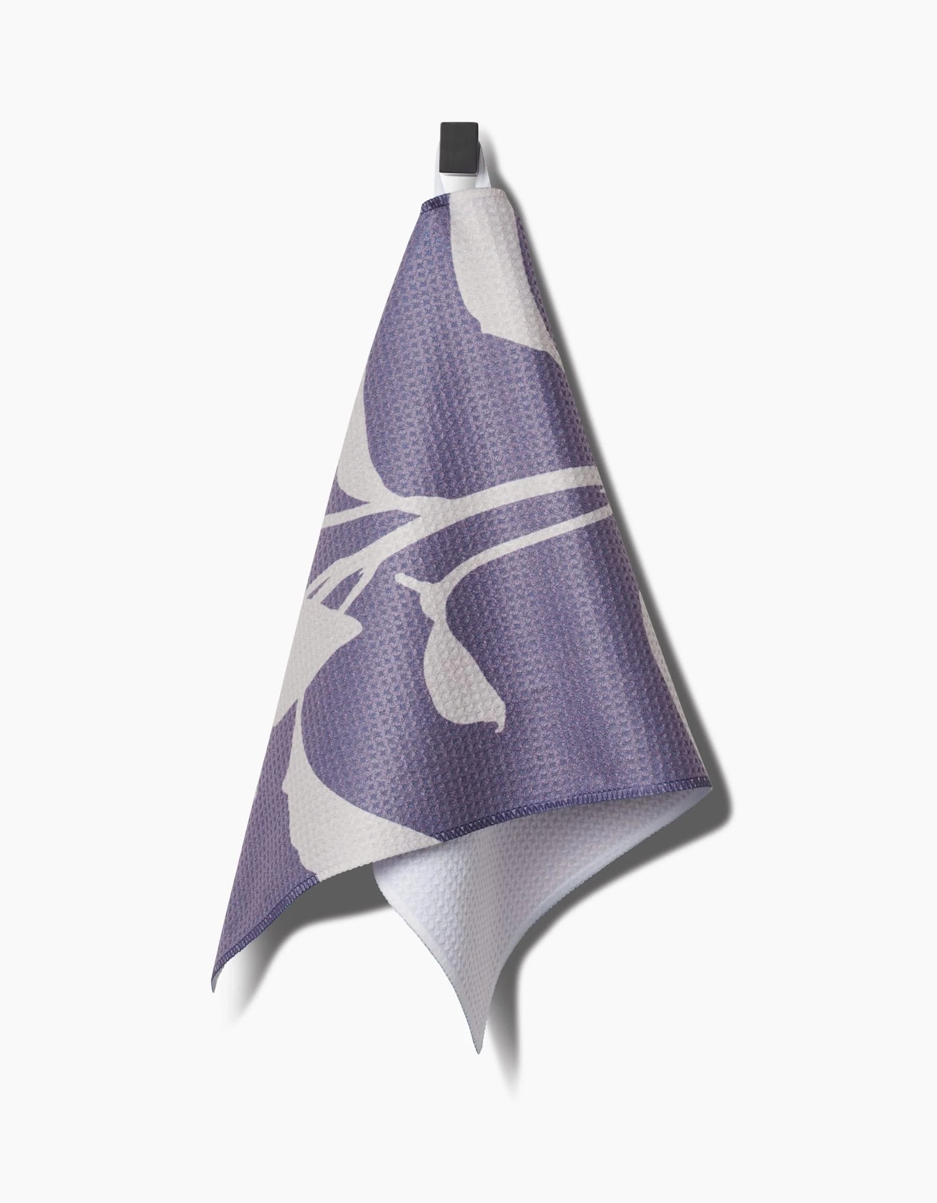 A Pepper bar towel by PrintedPlant, featuring a bold purple and white abstract floral design made from recycled materials, hangs on a black hook against a plain white background.