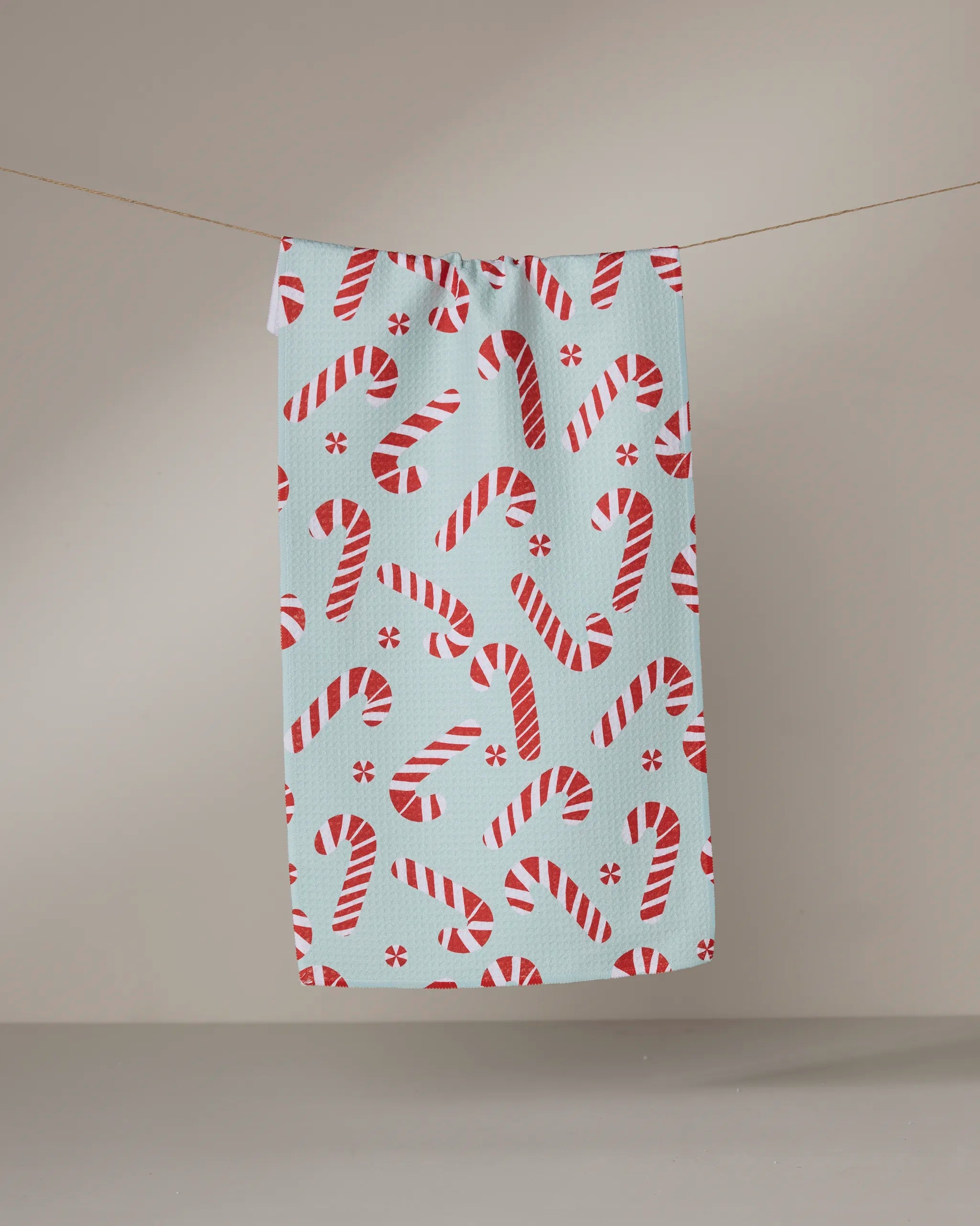 The Peppermint Twist towel by Geometry, in light blue with a red and white candy cane pattern, dries quickly and hangs on a thin clothesline against a plain beige background.