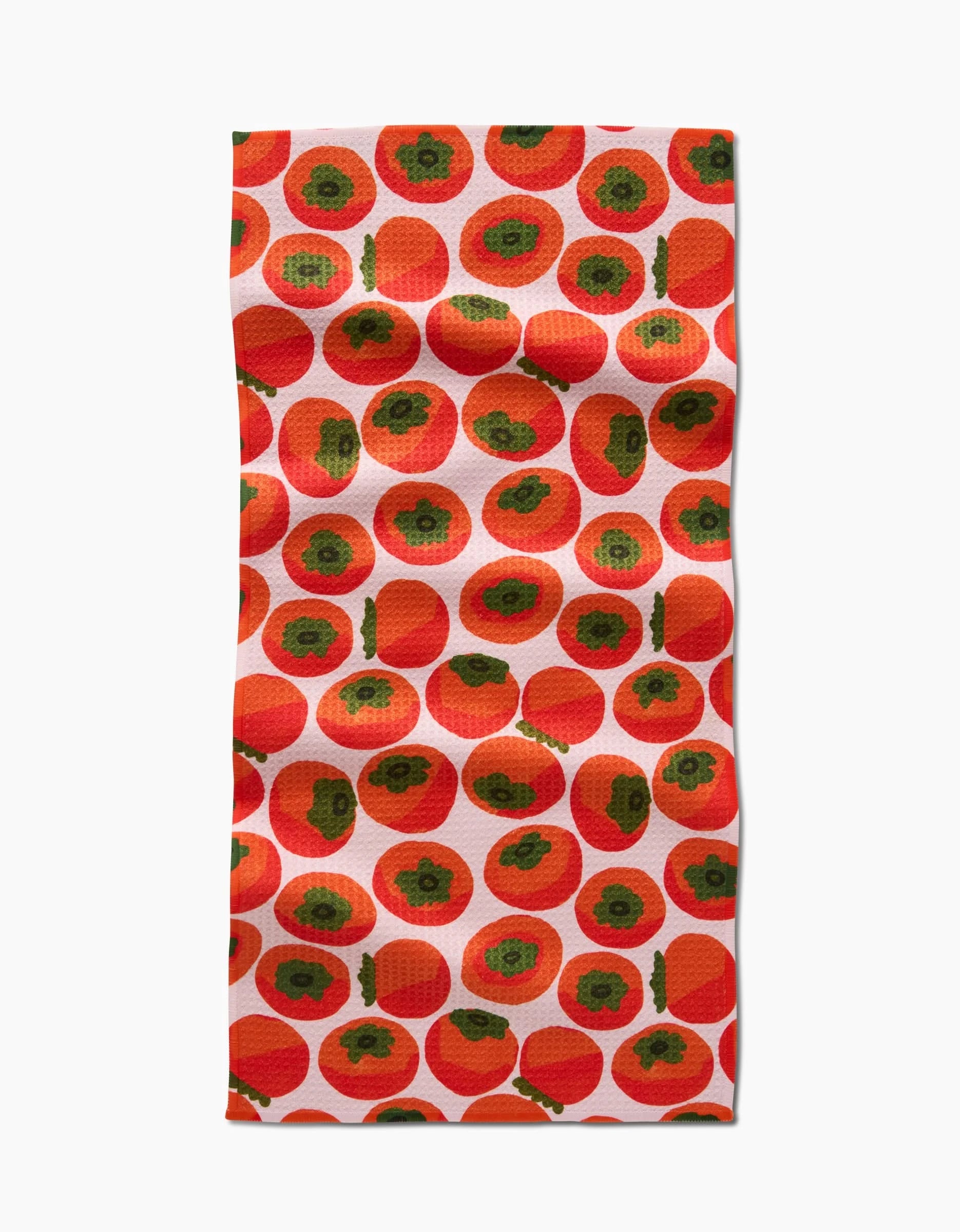 A rectangular bar towel by Katie OShea, made from Geoweave fabric, showcases a repeating pattern of vibrant persimmons with dark seeds and green stems on a white background.