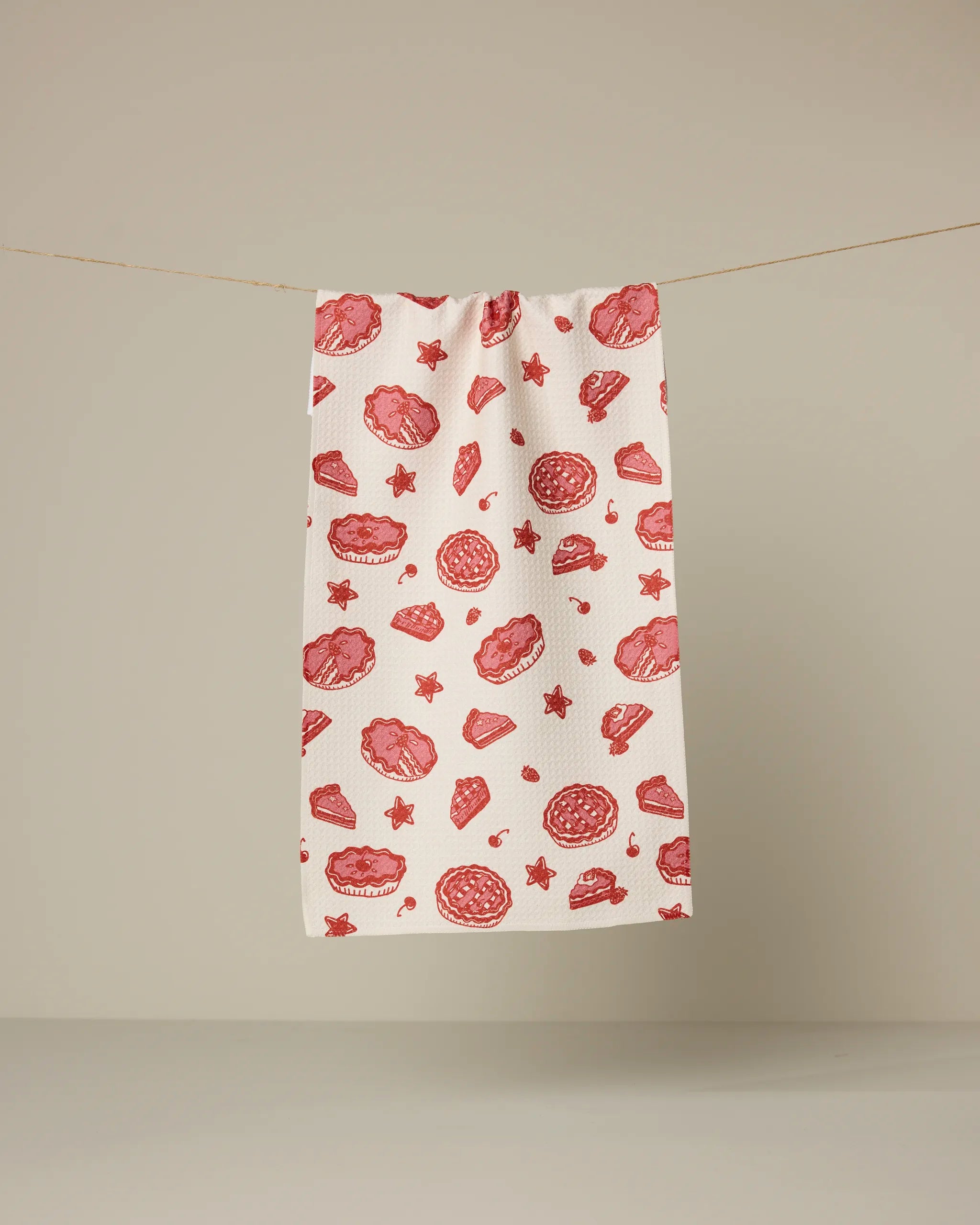 Geometry’s Pie-triotic recycled kitchen towel, featuring red illustrations of cookies, stars, and baked goods, hangs on a string against a plain beige background.