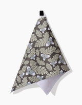 The Pinecone Patch kitchen towel by Danelys Sidron features beige leaves and white acorns on a dark background, hangs from a black loop against a white surface, and has a white textured back.