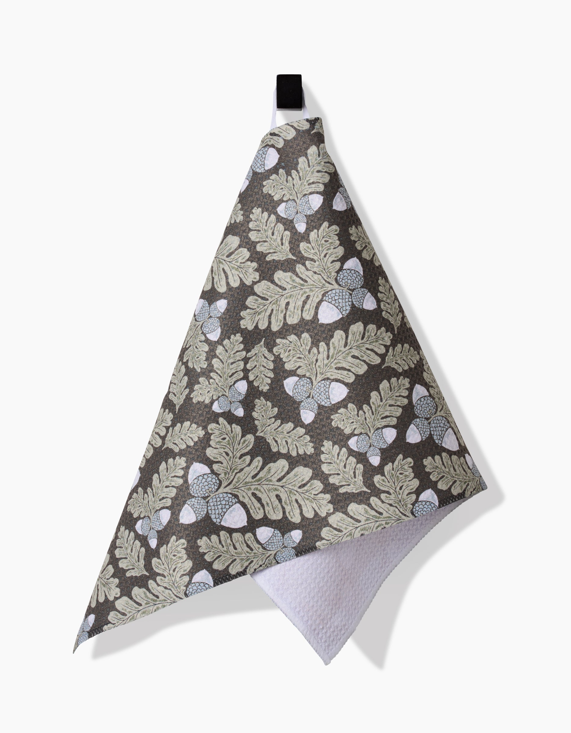 The Pinecone Patch kitchen towel by Danelys Sidron features beige leaves and white acorns on a dark background, hangs from a black loop against a white surface, and has a white textured back.