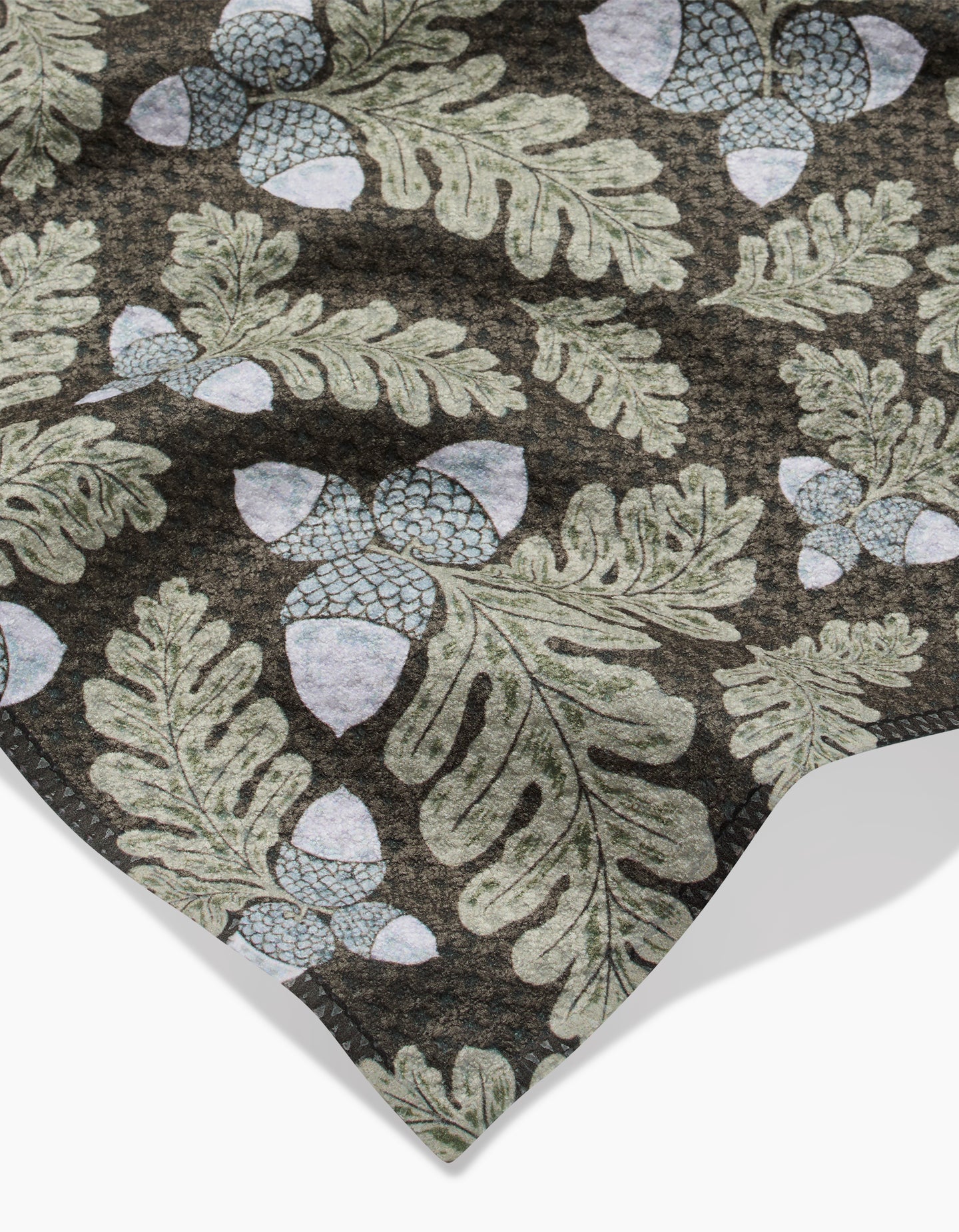 A close-up of Danelys Sidrons Pinecone Patch shows fabric with green oak leaves and pale blue acorns on a dark background. Slightly wrinkled, the ultra-absorbent towel highlights its soft, textured appearance.