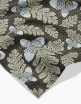 A close-up of Danelys Sidrons Pinecone Patch shows fabric with green oak leaves and pale blue acorns on a dark background. Slightly wrinkled, the ultra-absorbent towel highlights its soft, textured appearance.