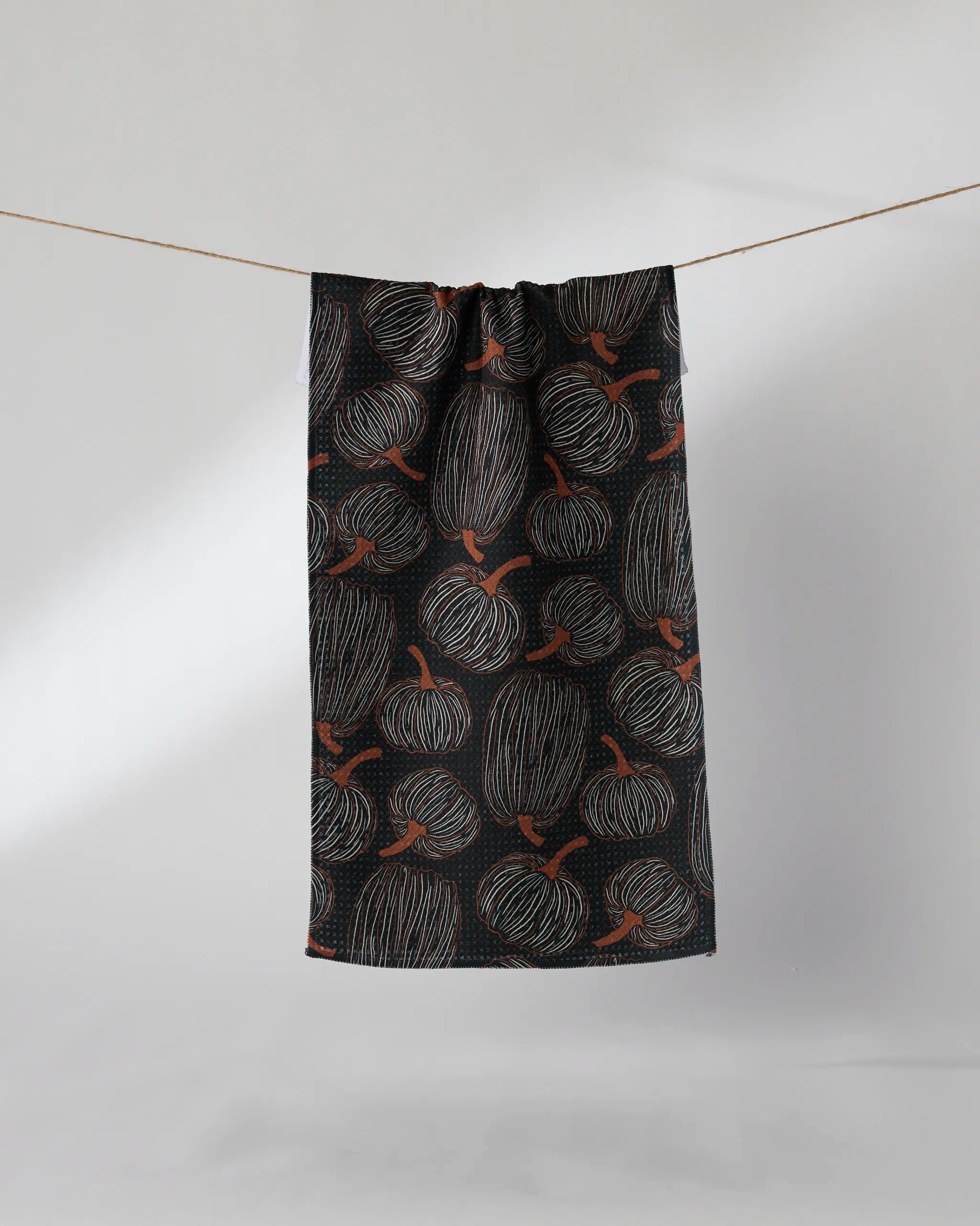 The Geometry Pinstripe Patch is a black ultra-absorbent towel featuring a repeating pattern of orange pumpkins outlined in white, shown hanging on a clothesline against a plain light background.