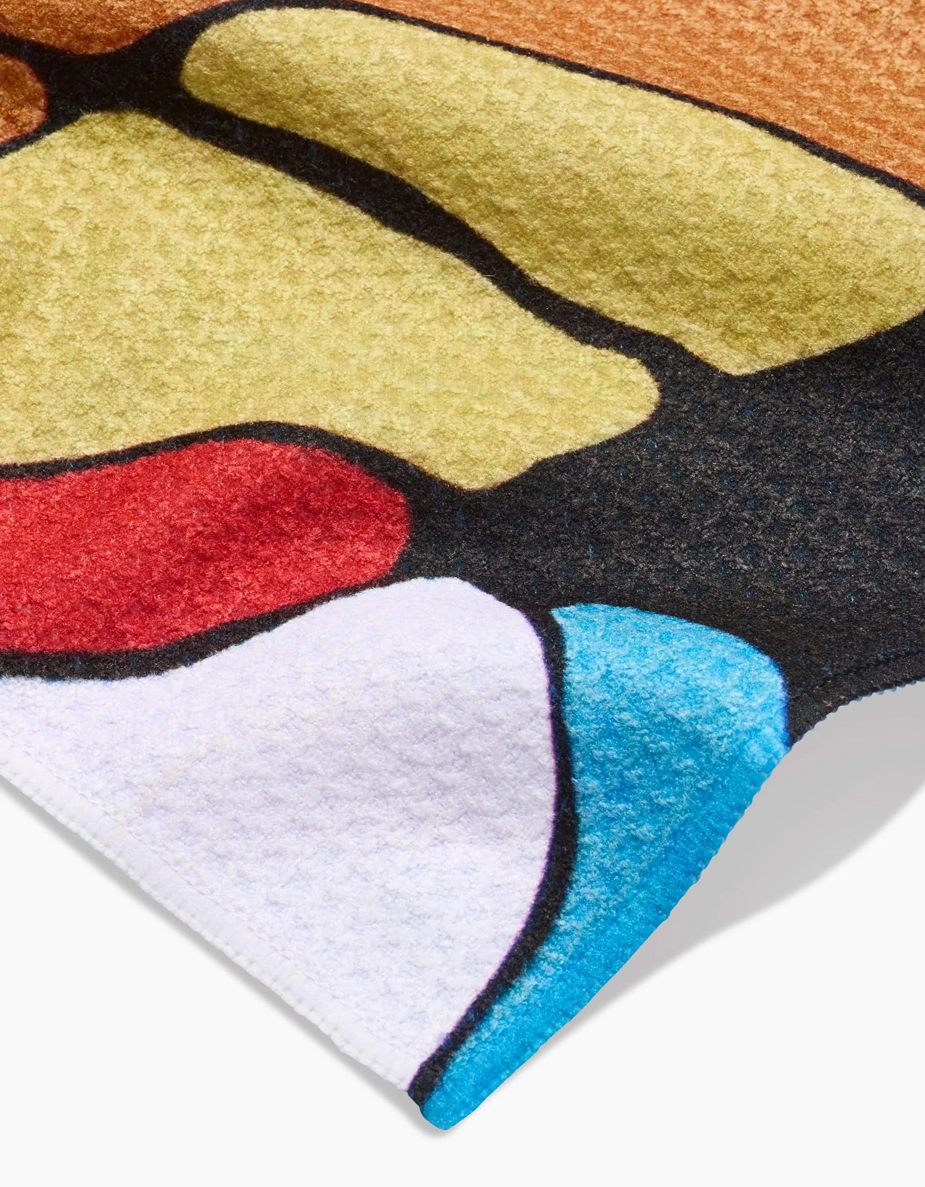 A close-up of the Rachel Austin Poppies on Blue Bar Towel shows its colorful, textured Geoweave fabric with bold shapes in yellow, red, black, white, blue, and brown. The soft towel’s corner curls slightly.