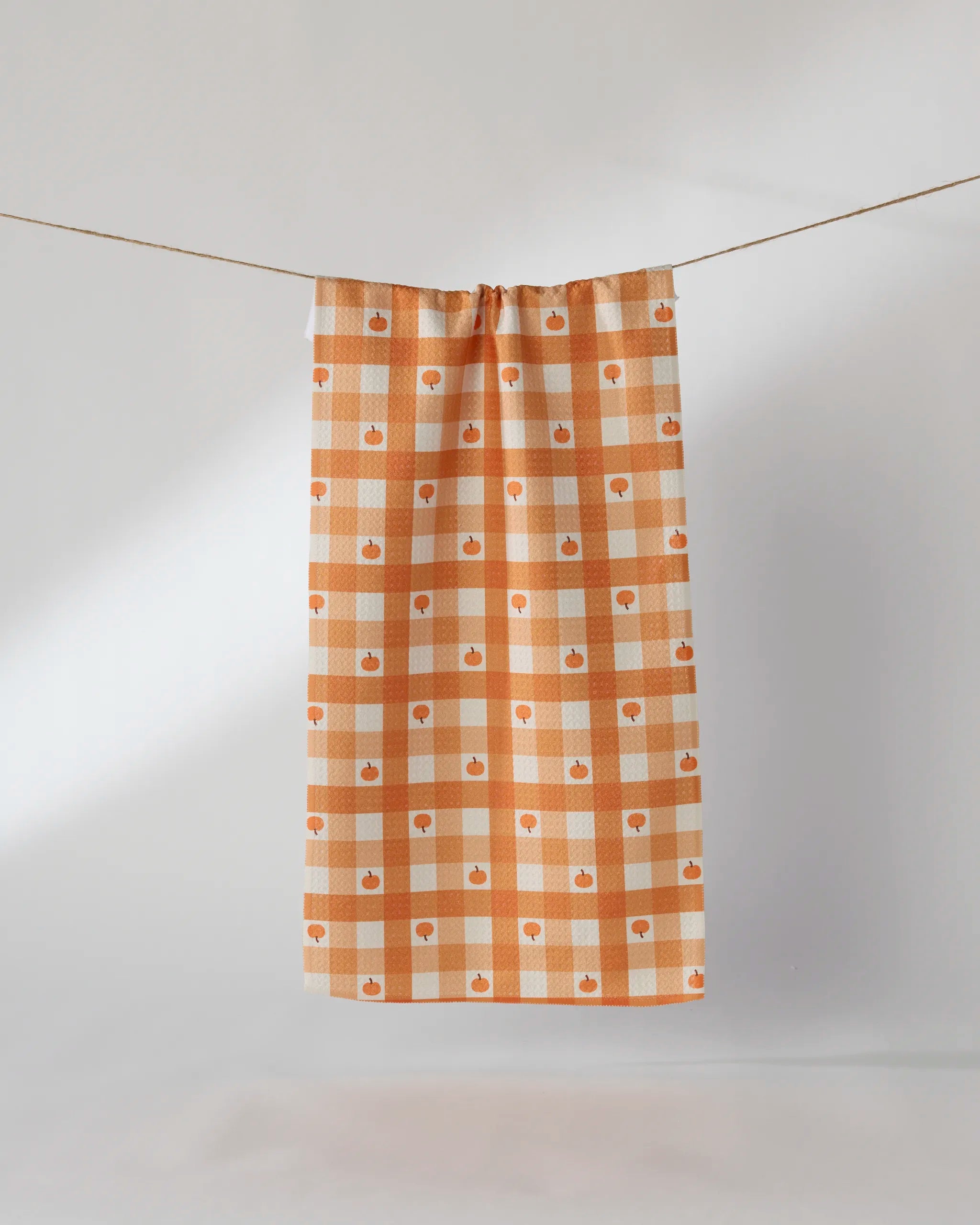 The Geometry Pumpkin Check bar towel, featuring an orange and white gingham design with a small apple pattern and made from recycled materials, hangs on a clothesline against a simple light background.
