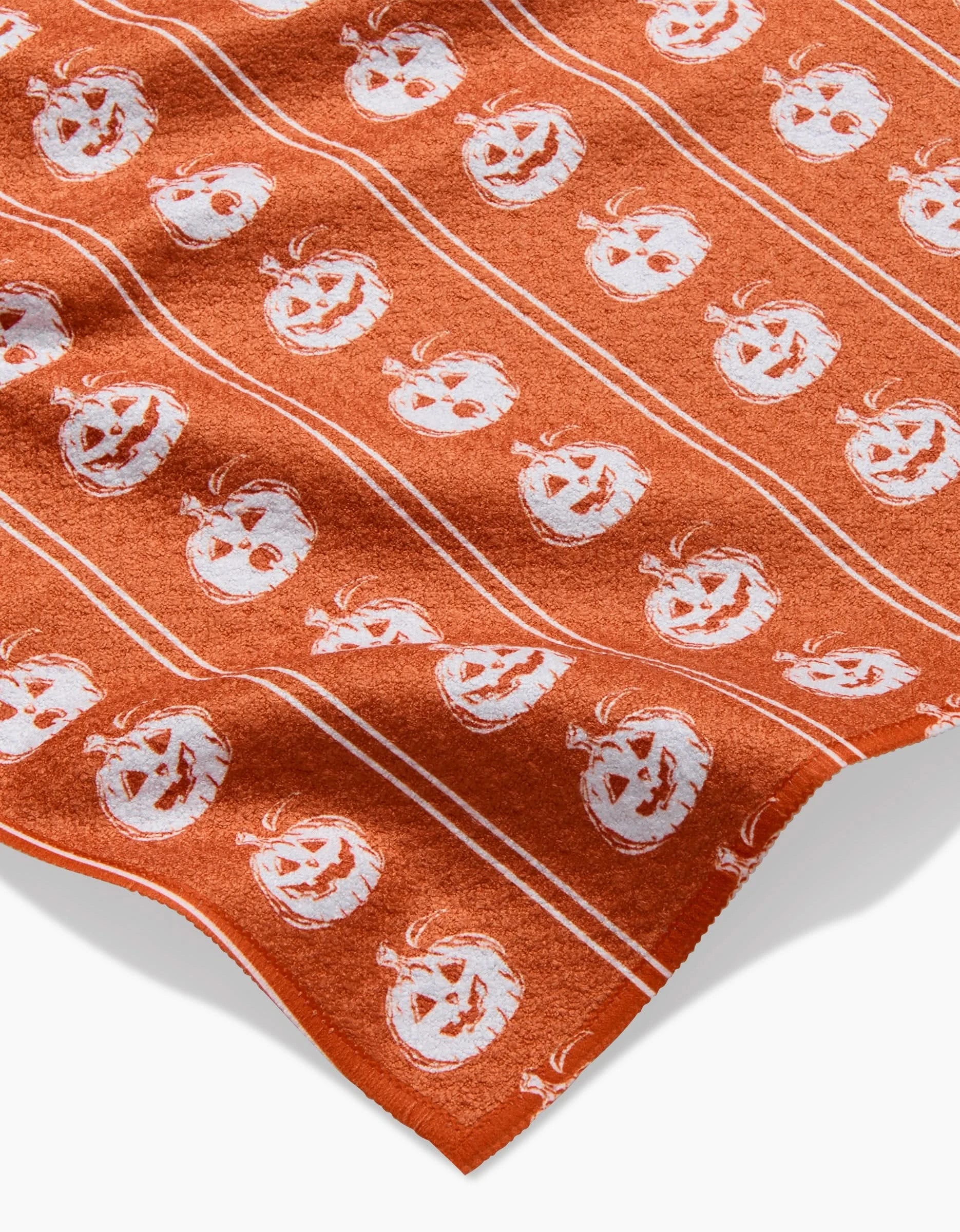 The Pumpkin Pinstripes bar towel by Aubrey Fairchild features white jack-o’-lantern faces and thin stripes in a repeating pattern on orange. Ultra-absorbent, it has a slightly wrinkled look and is shown against a light background.