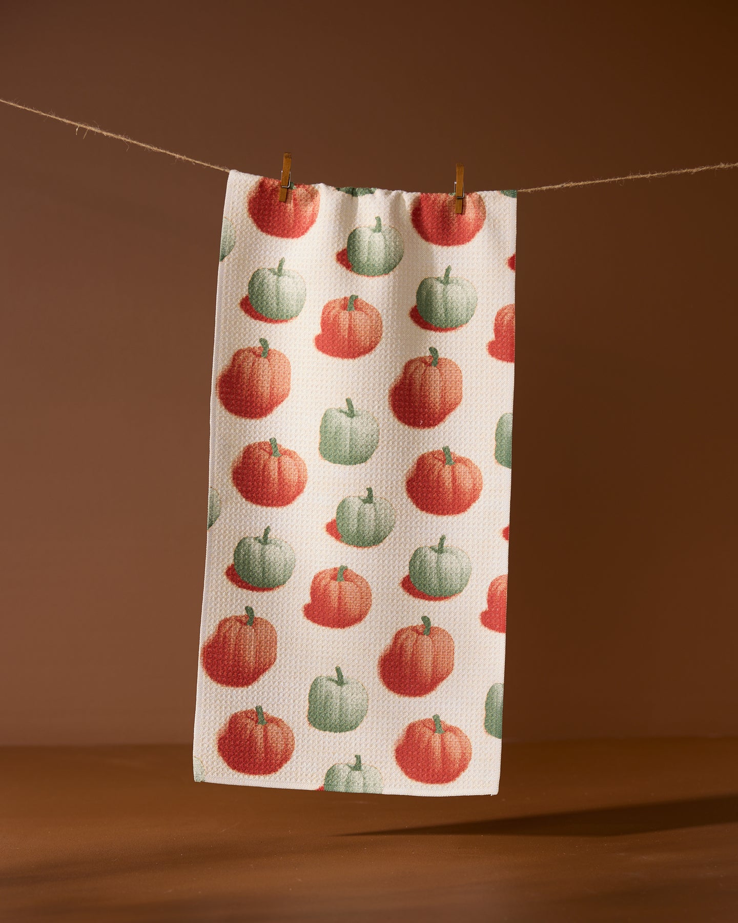 The Geometry Pumpkin Pop towel, ultra-absorbent and white with green and orange pumpkin patterns, hangs by two clothespins on a string against a brown background.