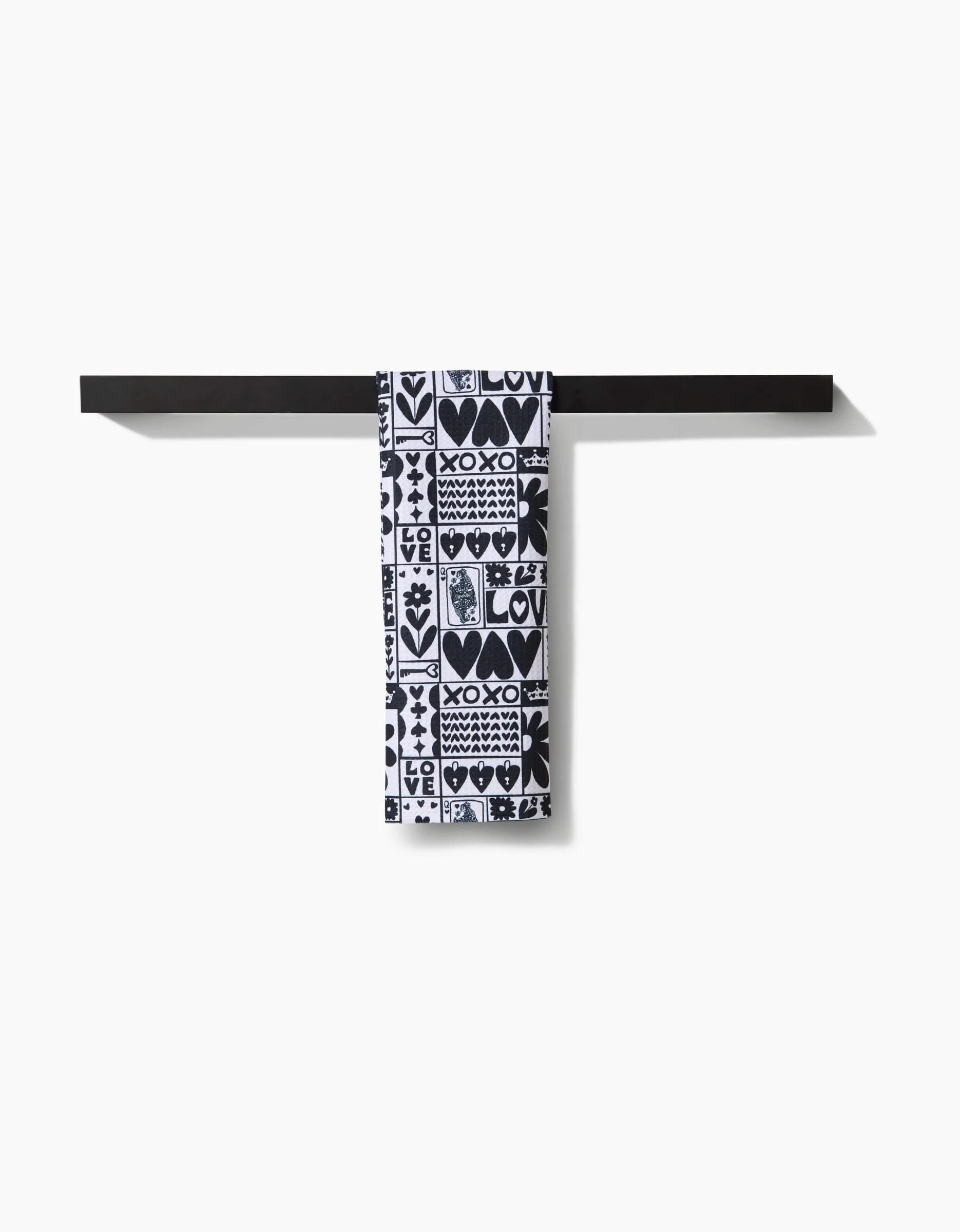 The Kayla Ann Queen of Hearts bar towel, featuring black and white designs with hearts and “LOVE,” hangs neatly on a black towel bar against a plain white background.