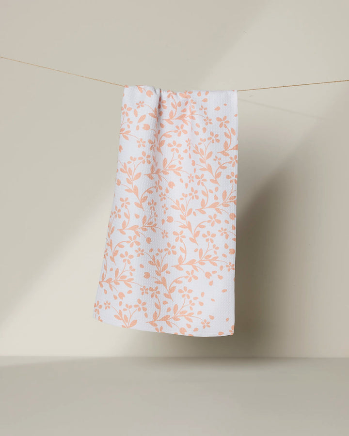 The Geometry Quiet Floral towel, featuring a peach floral pattern on white, hangs from a thin string against a neutral light background.