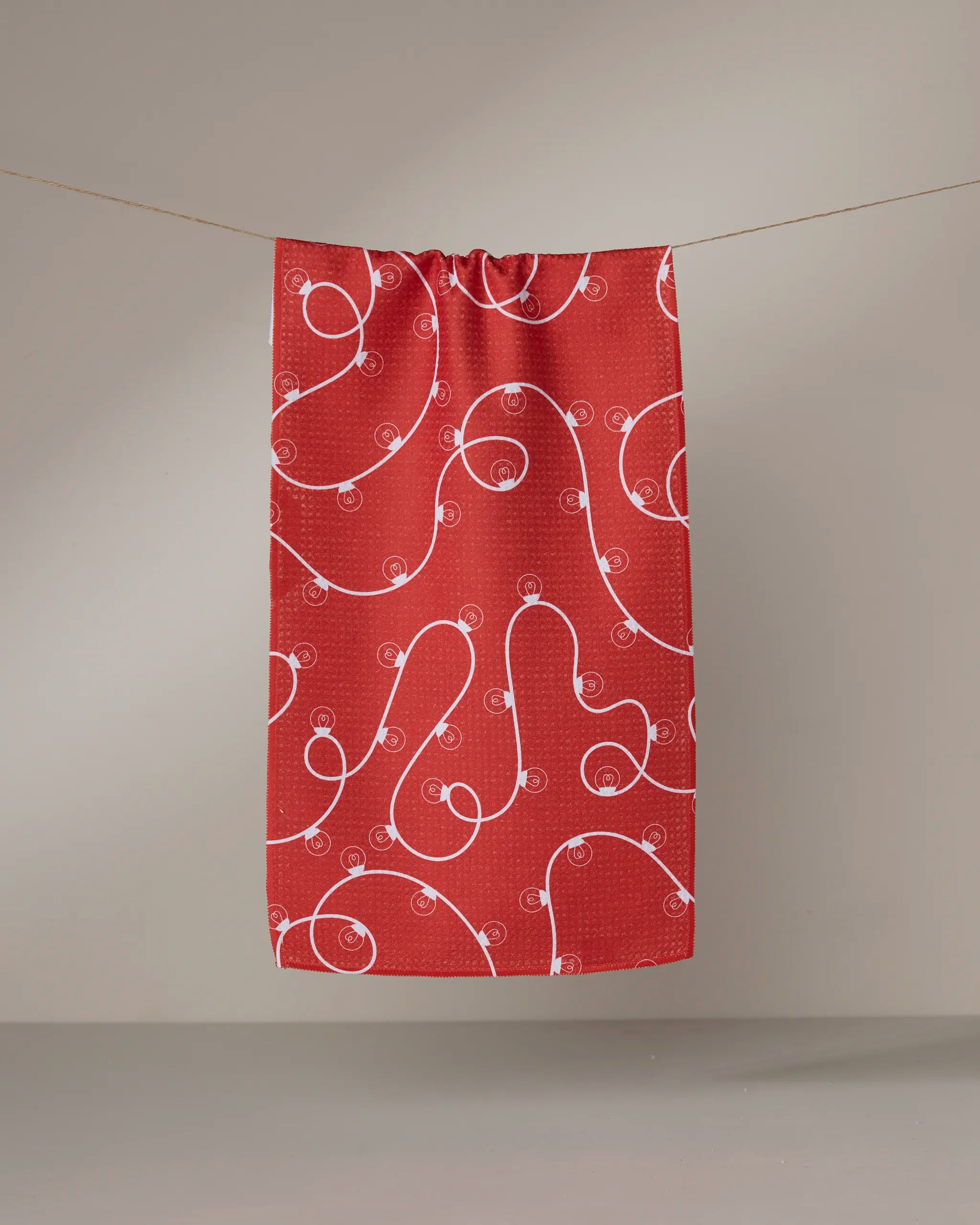 The Geometry Red Holiday Lights towel, featuring a quick-dry fabric and an abstract white swirl and dot design, hangs neatly on a clothesline against a plain gray background.