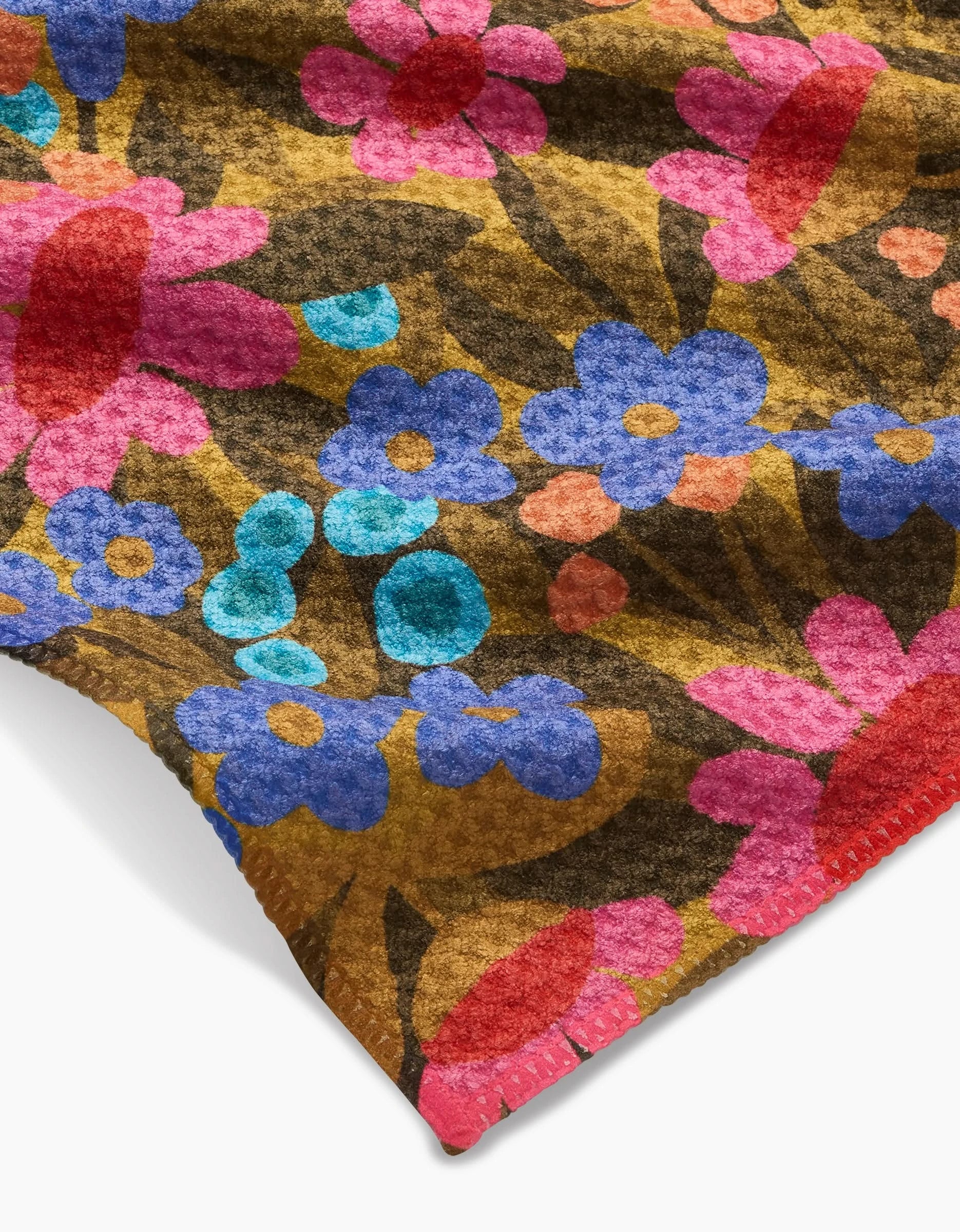 A close-up of the Lisa Nelson Retro Garden Bar Towel, featuring a vibrant floral print with blue, pink, and red flowers on a brown background. Made from Geoweave fabric, the towel’s edges are visible in the foreground.