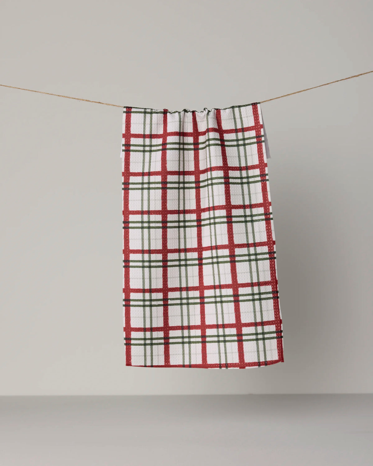 A Geometry Ribbon Plaid mini towel, featuring a red and green plaid pattern on white Geoweave fabric, hangs from a string against a plain gray background.