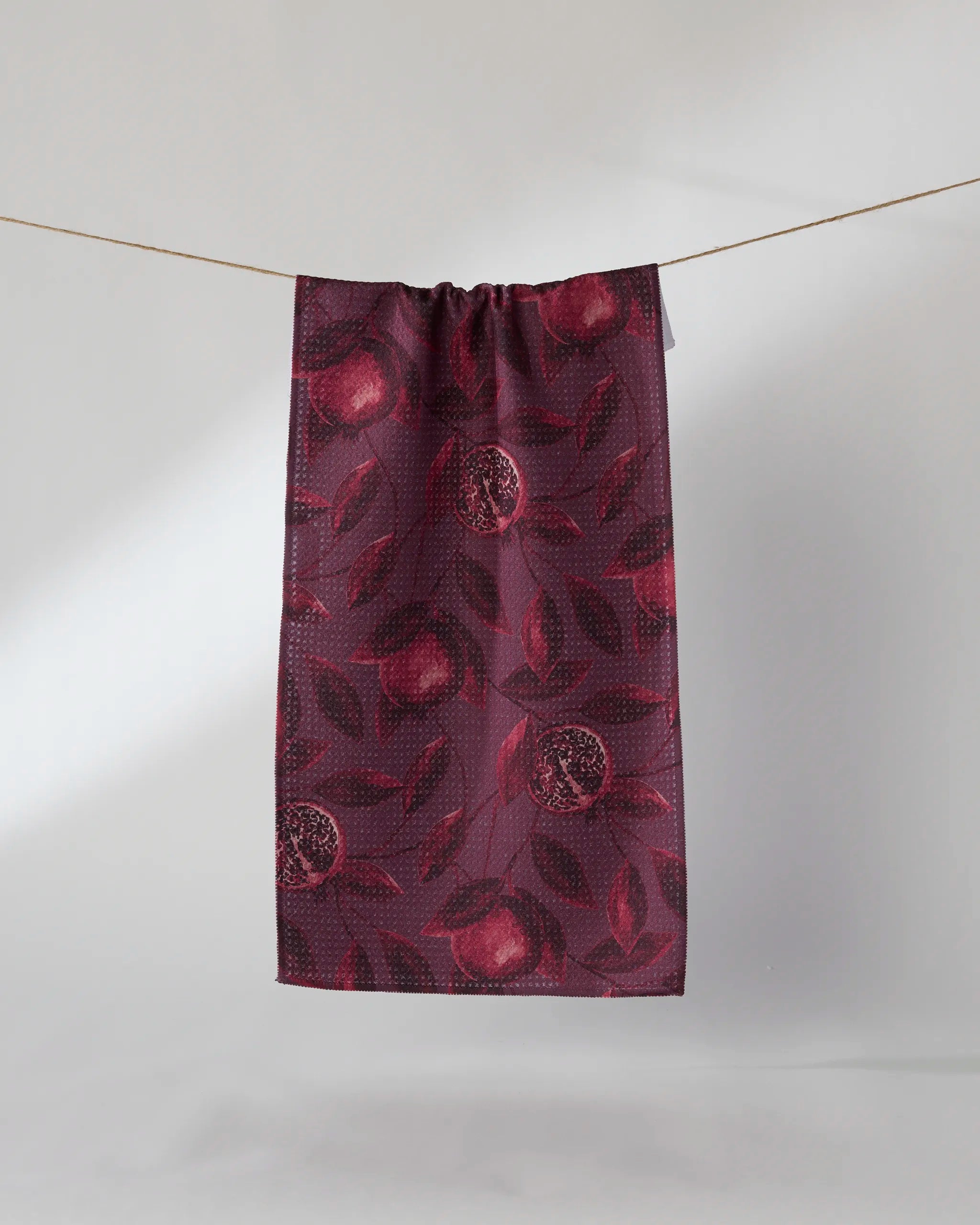 The Geometry "Rich Garnet" tea towel, featuring a pomegranate and leaf motif in dark red, hangs from a thin string against a light gray background.