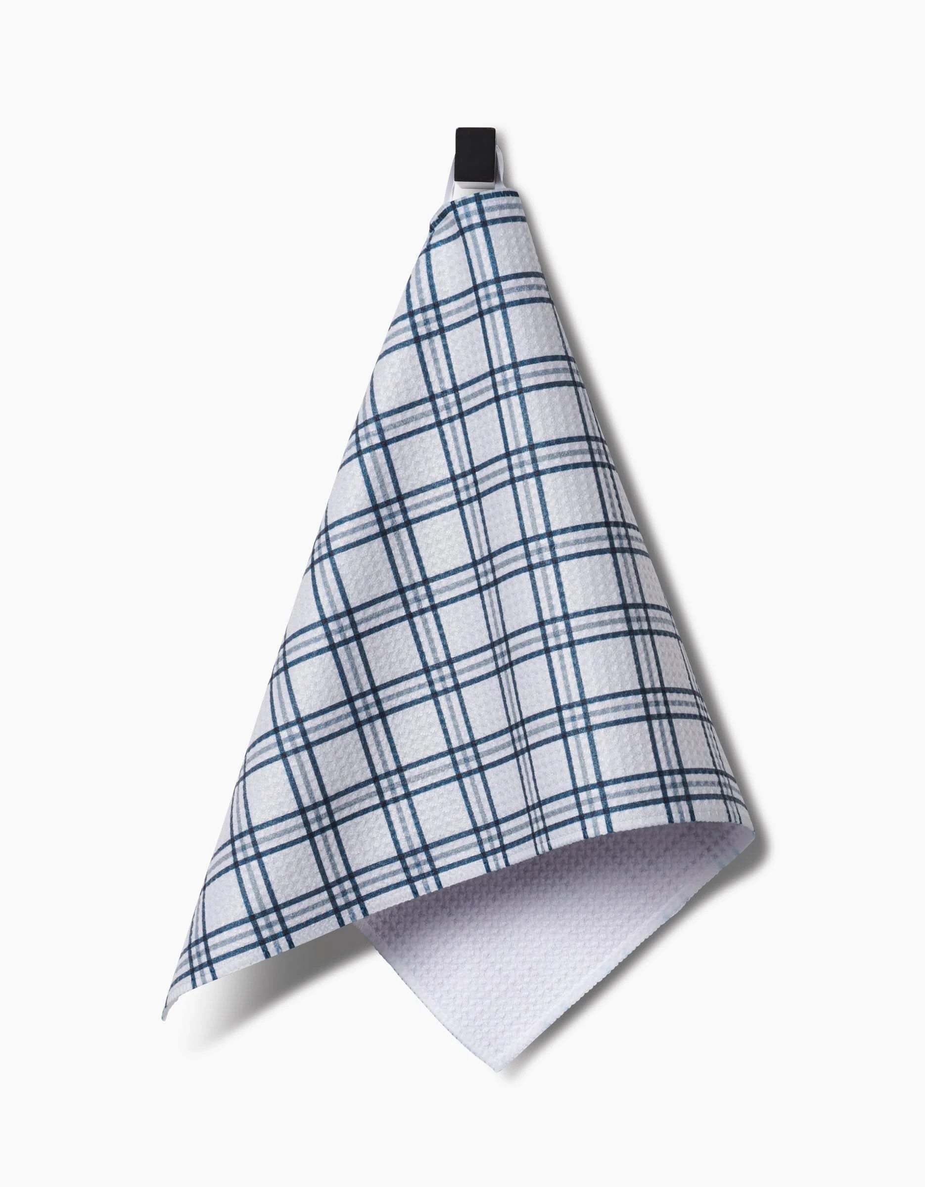The Roma Bar Towel by Kelsi Fullmer, featuring a blue plaid pattern on white Geoweave fabric, hangs from a black loop against a plain white background.