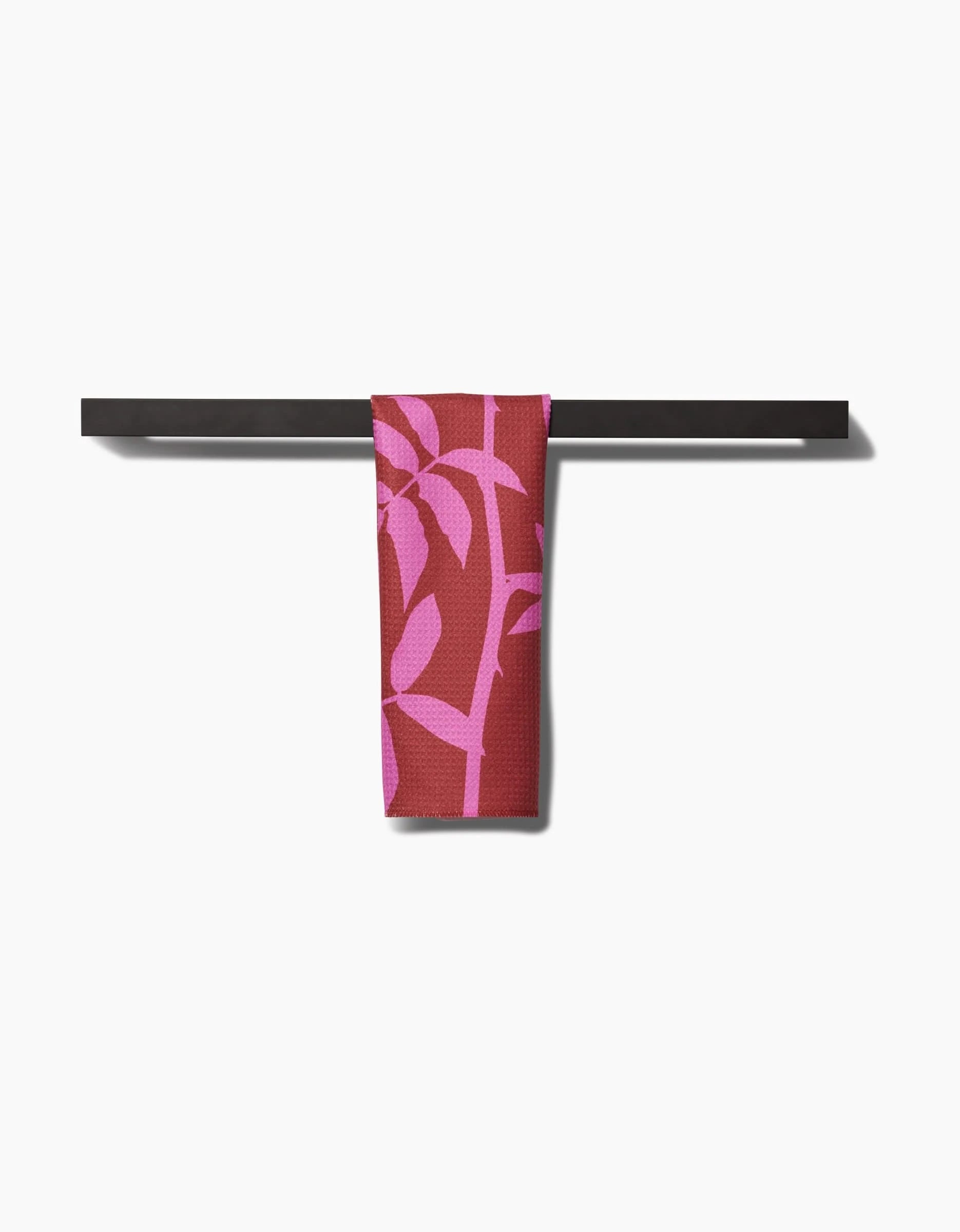 The PrintedPlant Rose bar towel, featuring a pink leaf design on maroon and made from recycled materials, hangs over a black towel bar against a white background.