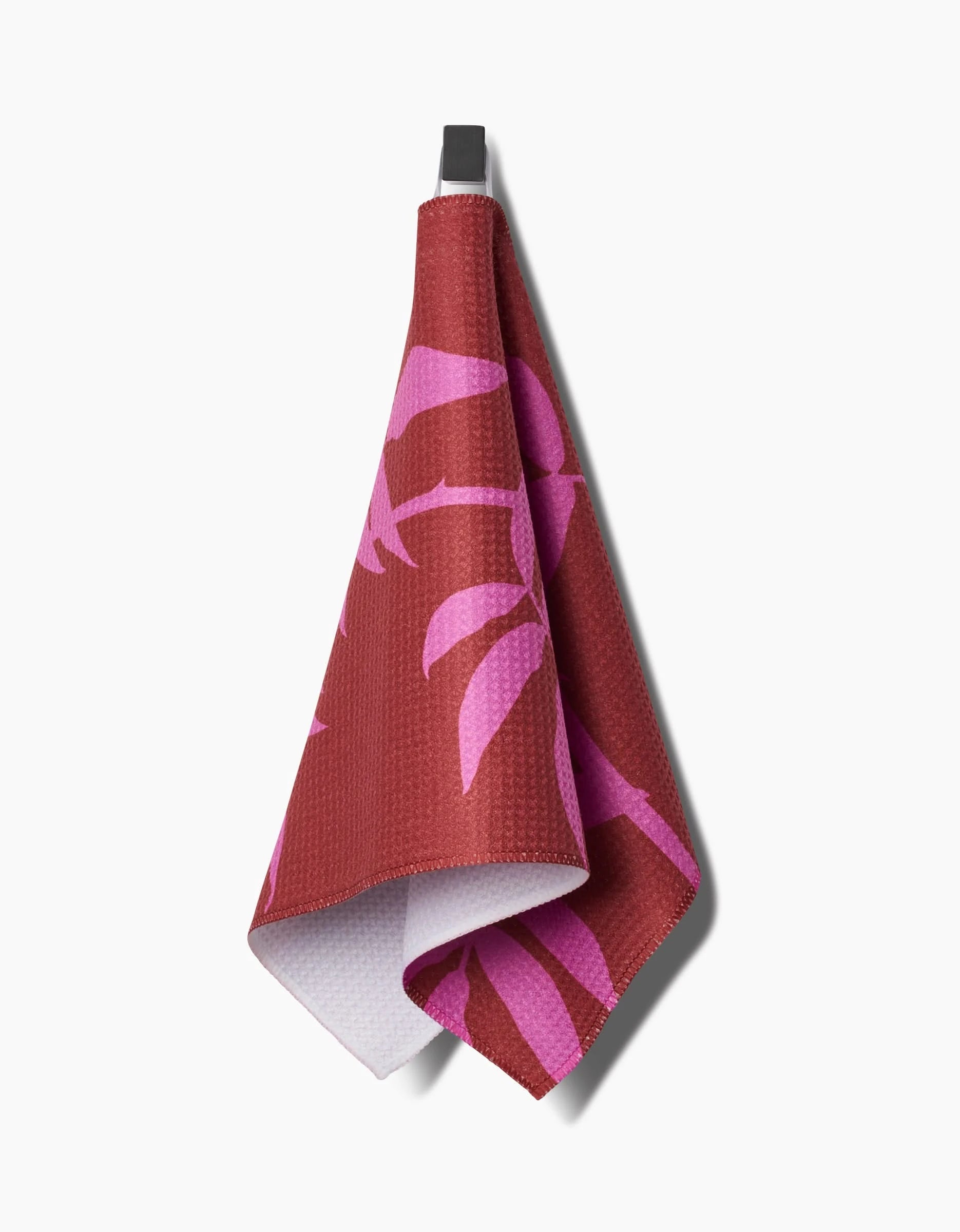 The PrintedPlant Rose Bar Towel features maroon and pink palm leaf patterns on Geoweave fabric, a textured front, a white back, and hangs from a black hook against a white background.