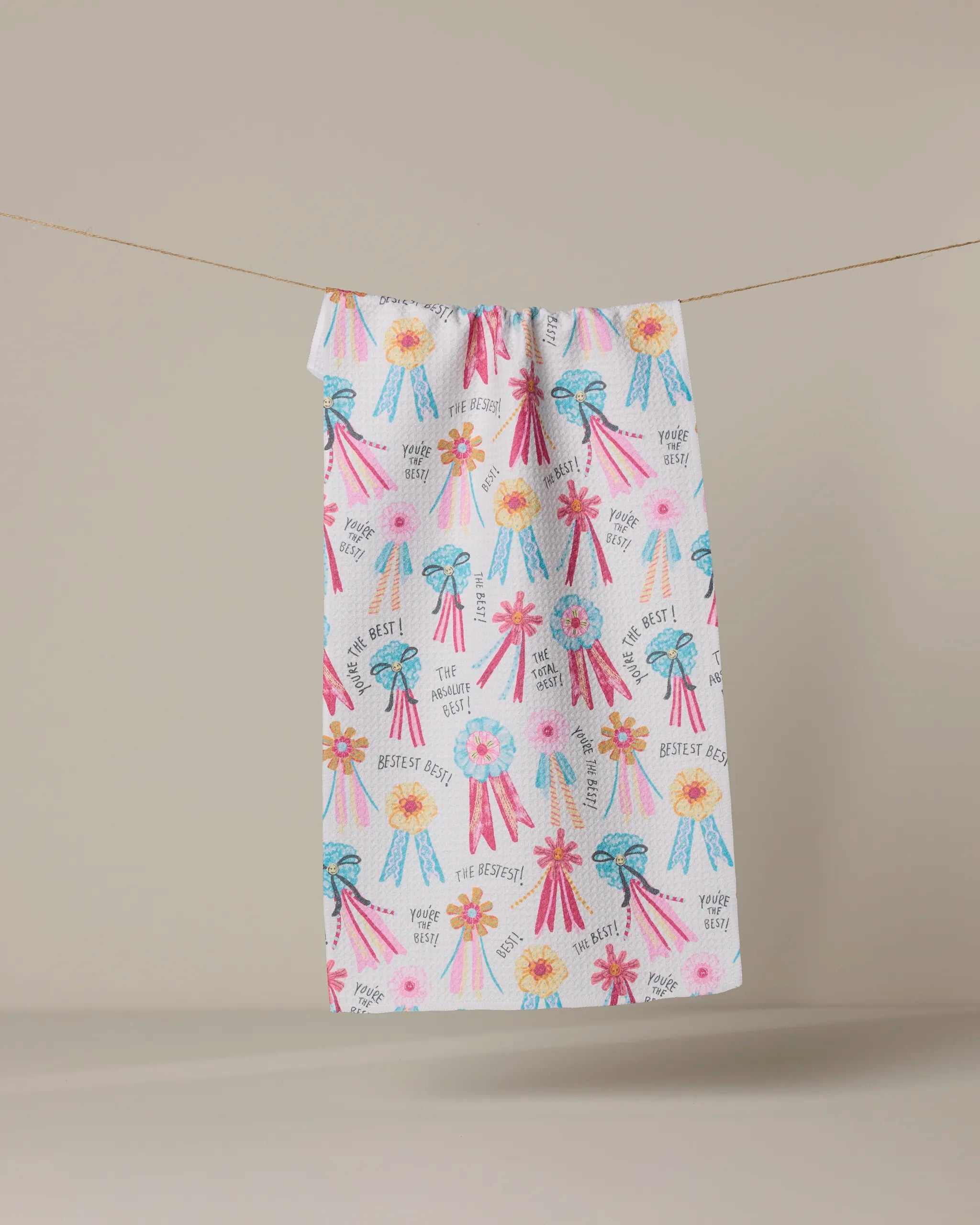 The Geometry "Rosettes for You" towel is ultra-absorbent and colorful, adorned with ribbons, rosettes, and phrases like "Bestest Best" and "You're The Best!"—shown hanging on a clothesline against a beige background.