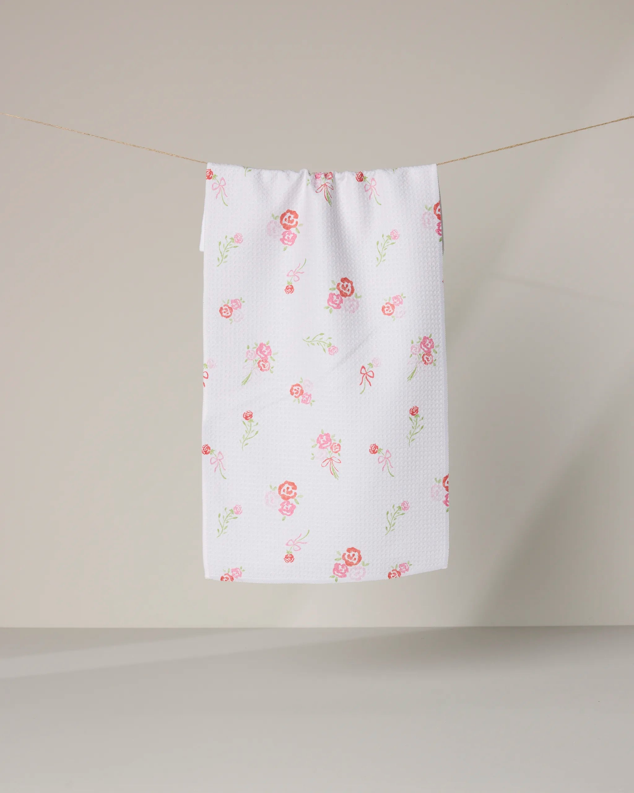 The Rosy Cheeks towel by Geometry, featuring a pink floral pattern and white textured ultra-absorbent fabric, hangs on a clothesline against a plain light beige background.