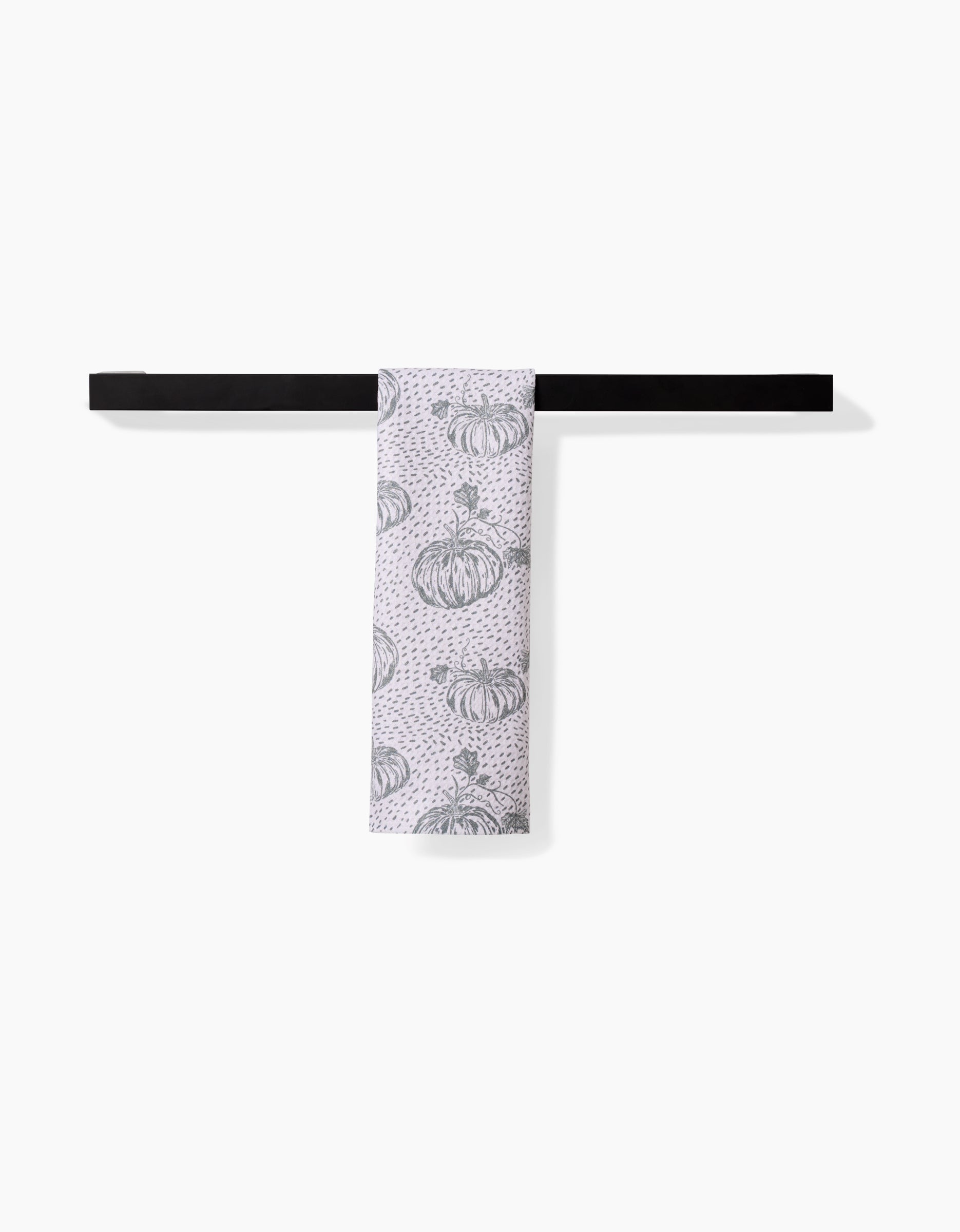The Sage Pumpkin Field bar towel by Charlie Rowan Designs, featuring a black pumpkin pattern on white and crafted from recycled materials, is neatly folded over a simple black bar against a plain white background.