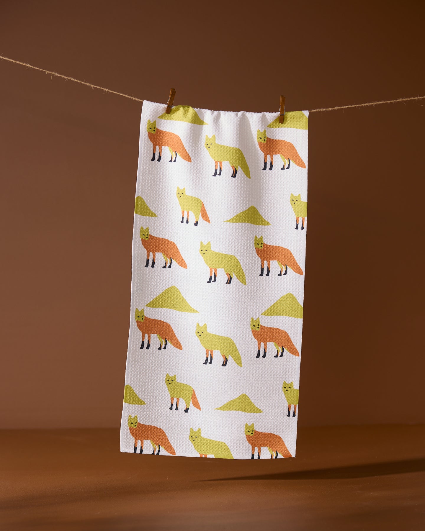 The Sagebrush Fox kitchen towel by Geometry, made from recycled materials, features orange and yellow foxes and green triangles in a repeating pattern. It hangs on a string with wooden clothespins against a brown background.