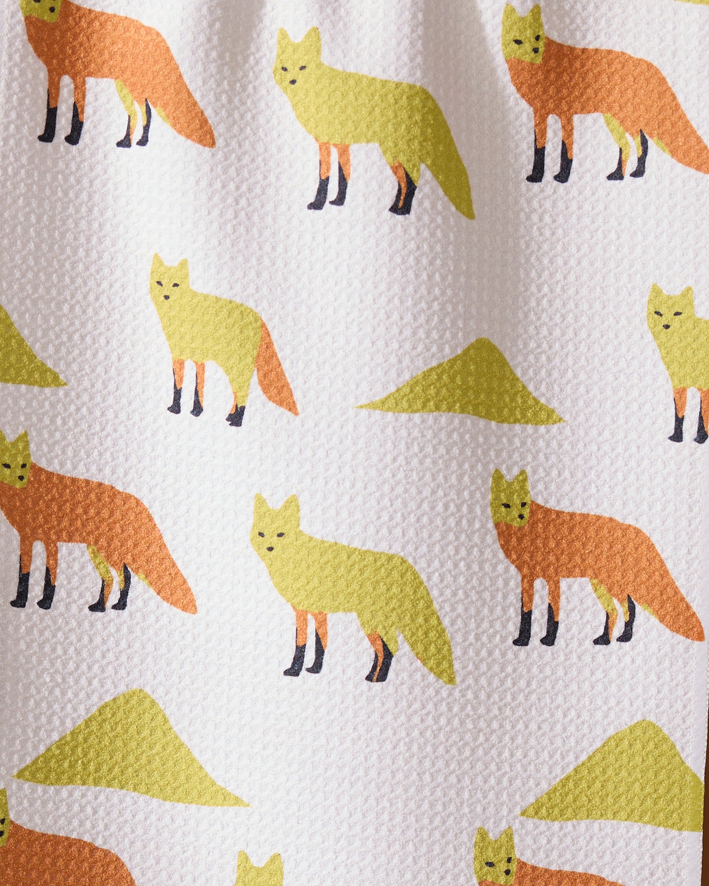 Geometry’s “Sagebrush Fox” kitchen towel features simple cartoon yellow and orange foxes with abstract green shapes on textured white fabric, thoughtfully crafted from recycled materials.